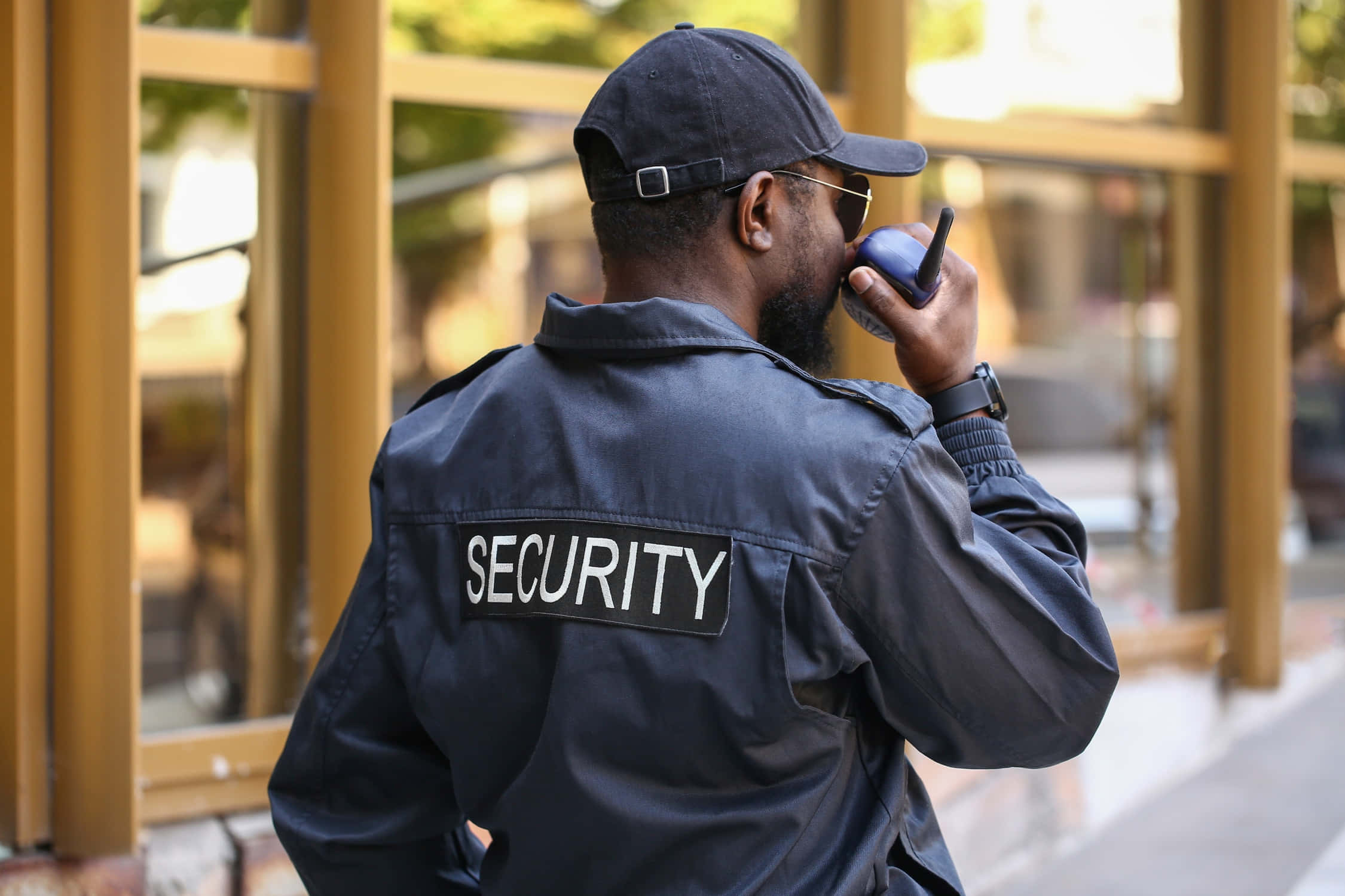 security 桌布 壁紙 — 一名身穿深色制服、背後印有「SECURITY」的保全人員在日光下於玻璃門附近使用對講機。