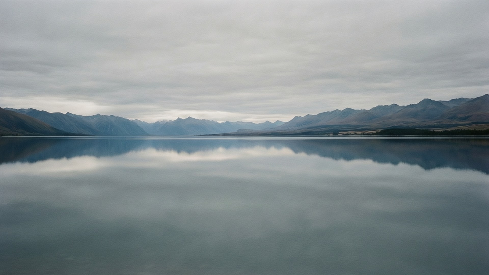Grey Lake Reflection 壁紙 — 一個寧靜的湖泊在陰天倒映著遠山，營造出對稱而寧靜的景觀。