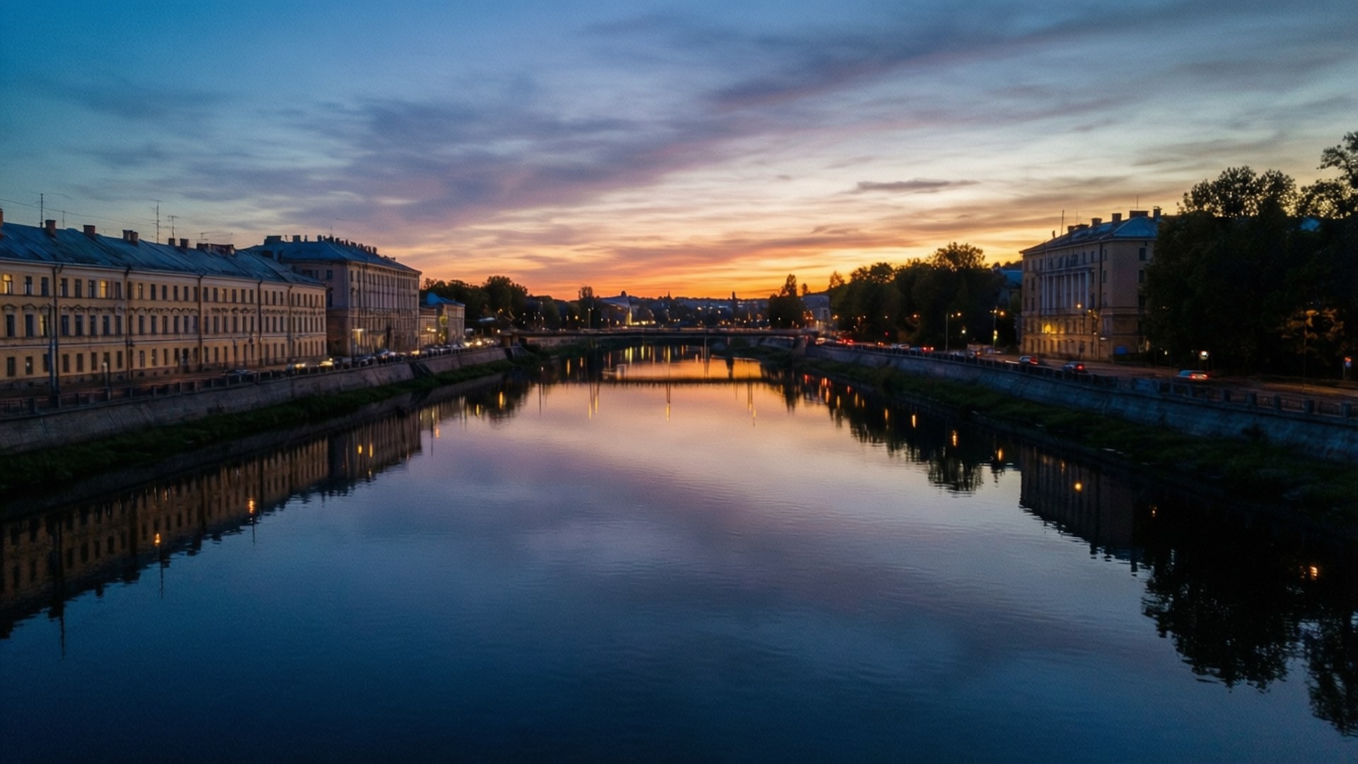 Neris River Dusk 壁紙 — 黃昏時分，一條寧靜的河流在歷史建築之間反射出溫暖的橙色和涼爽的藍色天空，建築物的窗戶閃爍著光芒。
