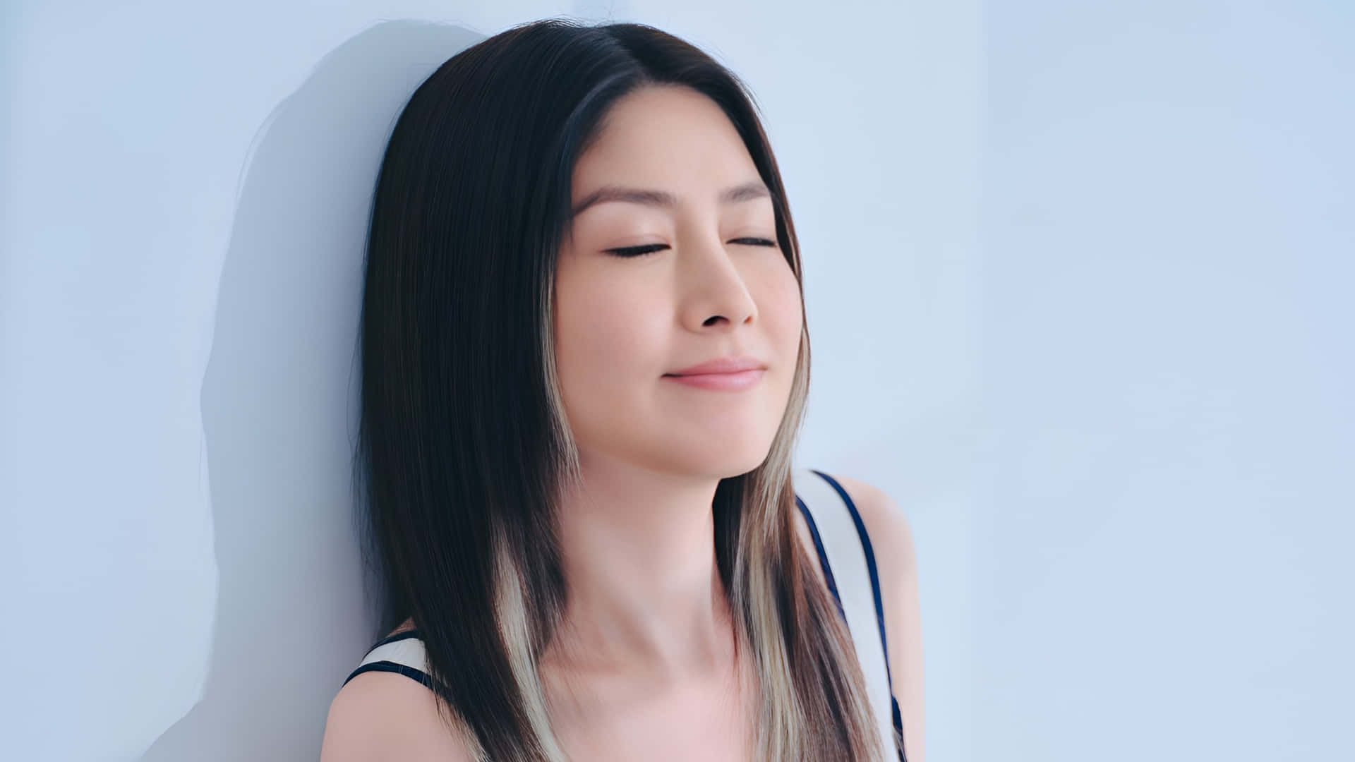  壁紙 — 一位深色頭髮的寧靜女子靠著淡藍色的牆壁，閉上眼睛，沉浸在平靜的滿足中。