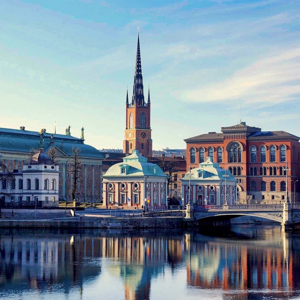 Linköping 桌布 壁紙 — 寧靜的城市景觀，歷史建築和高聳的教堂尖塔倒映在清澈藍天下的平靜水面上。