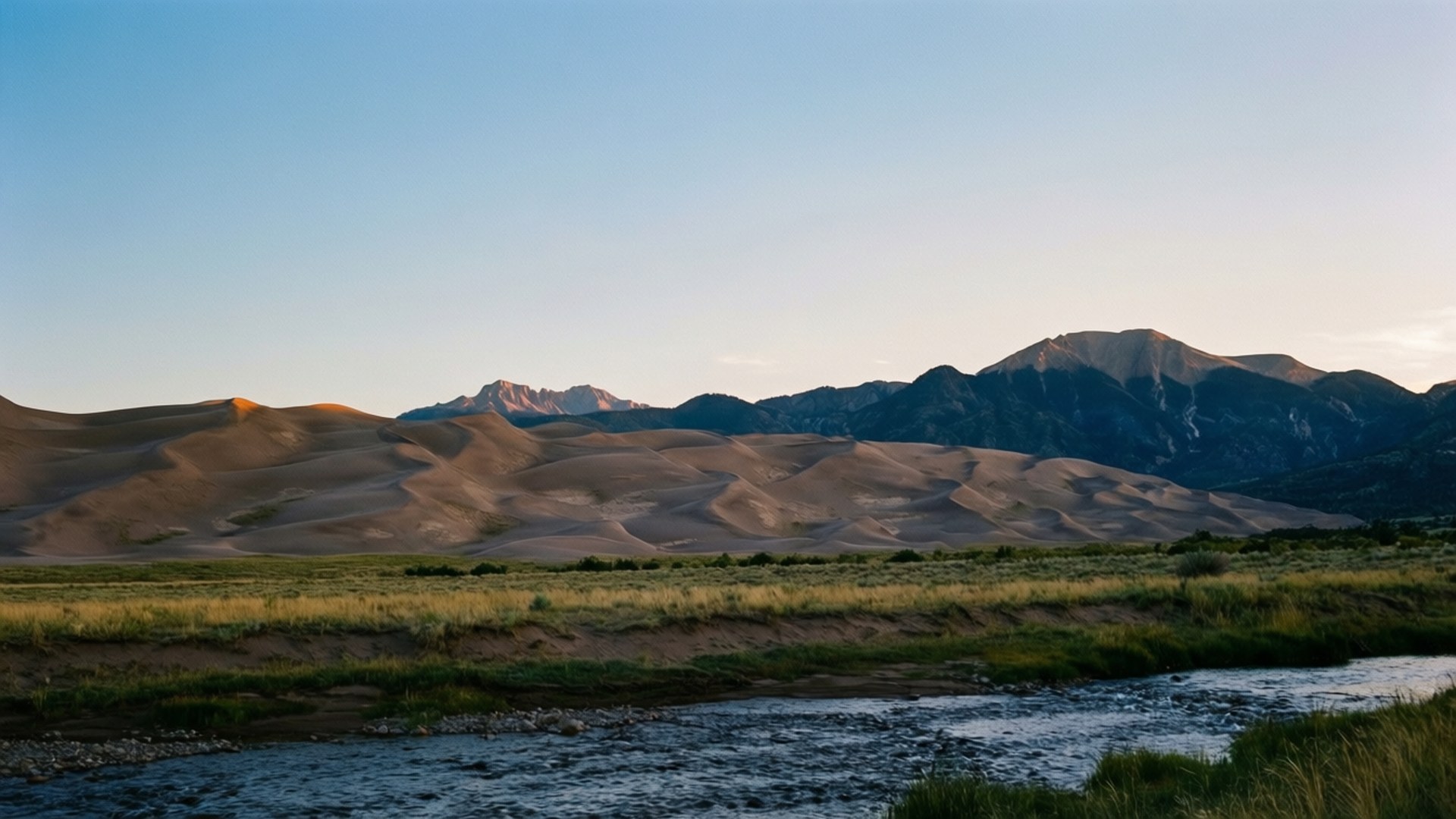 Great Sand Dunes Dusk 壁紙 — 寧靜的風景，前景是金色沙丘，蜿蜒的河流和遠處崎嶇的山脈，在清澈的藍天下。