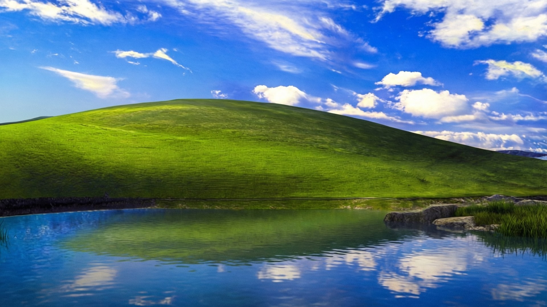 Bliss with River 壁紙 — 一個寧靜的風景，充滿活力的綠色山丘倒映在平靜的藍色湖泊中，天空明亮，有蓬鬆的白雲。