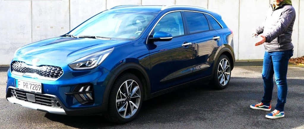 Kia Niro 桌布 壁紙 — 一輛閃亮藍色Kia Niro SUV停在柏油路上，旁邊有人做手勢，背景是樸素的混凝土牆。