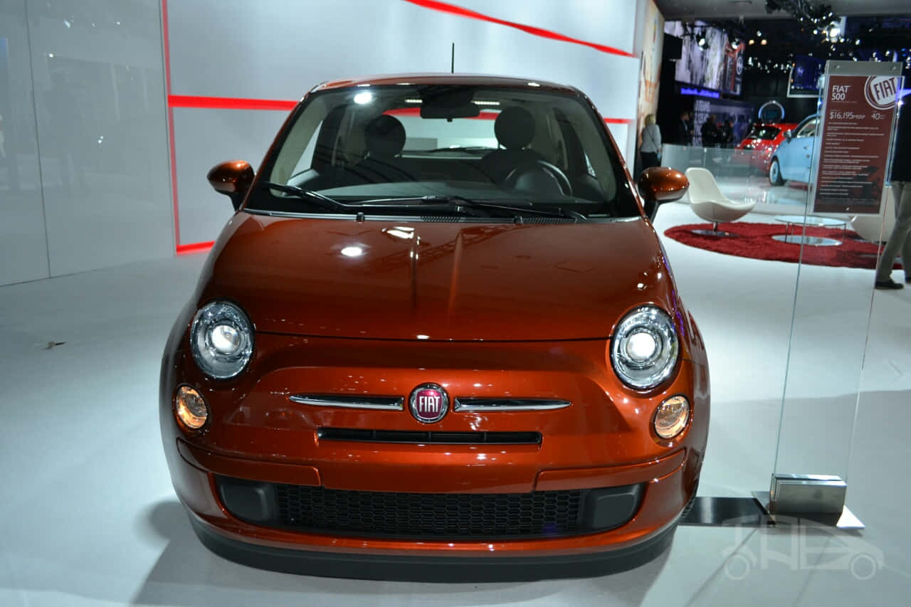 Fiat 500 桌布 壁紙 — 一輛閃亮的橙色Fiat 500在車展上展出，配有明亮的燈光和玻璃屏障。