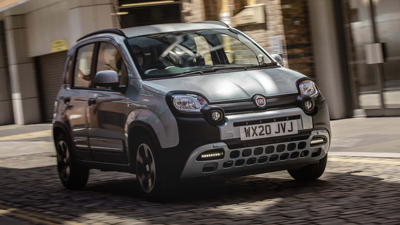 Fiat Panda 桌布 壁紙 — 一輛銀色的Fiat Panda在鵝卵石街道上行駛，背景是模糊的城市建築。