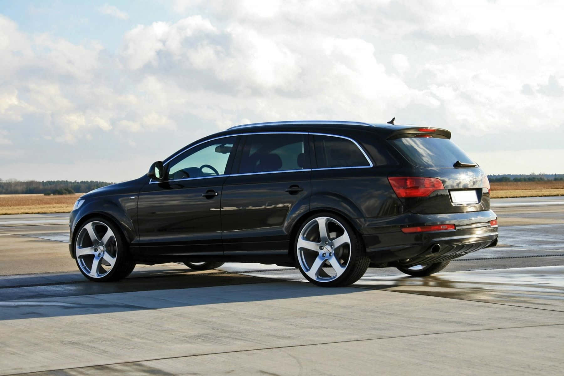 Audi Q7 桌布 壁紙 — 一輛時尚的黑色奧迪Q7 SUV停放在潮濕的停機坪上，天空多雲，遠處有樹木。