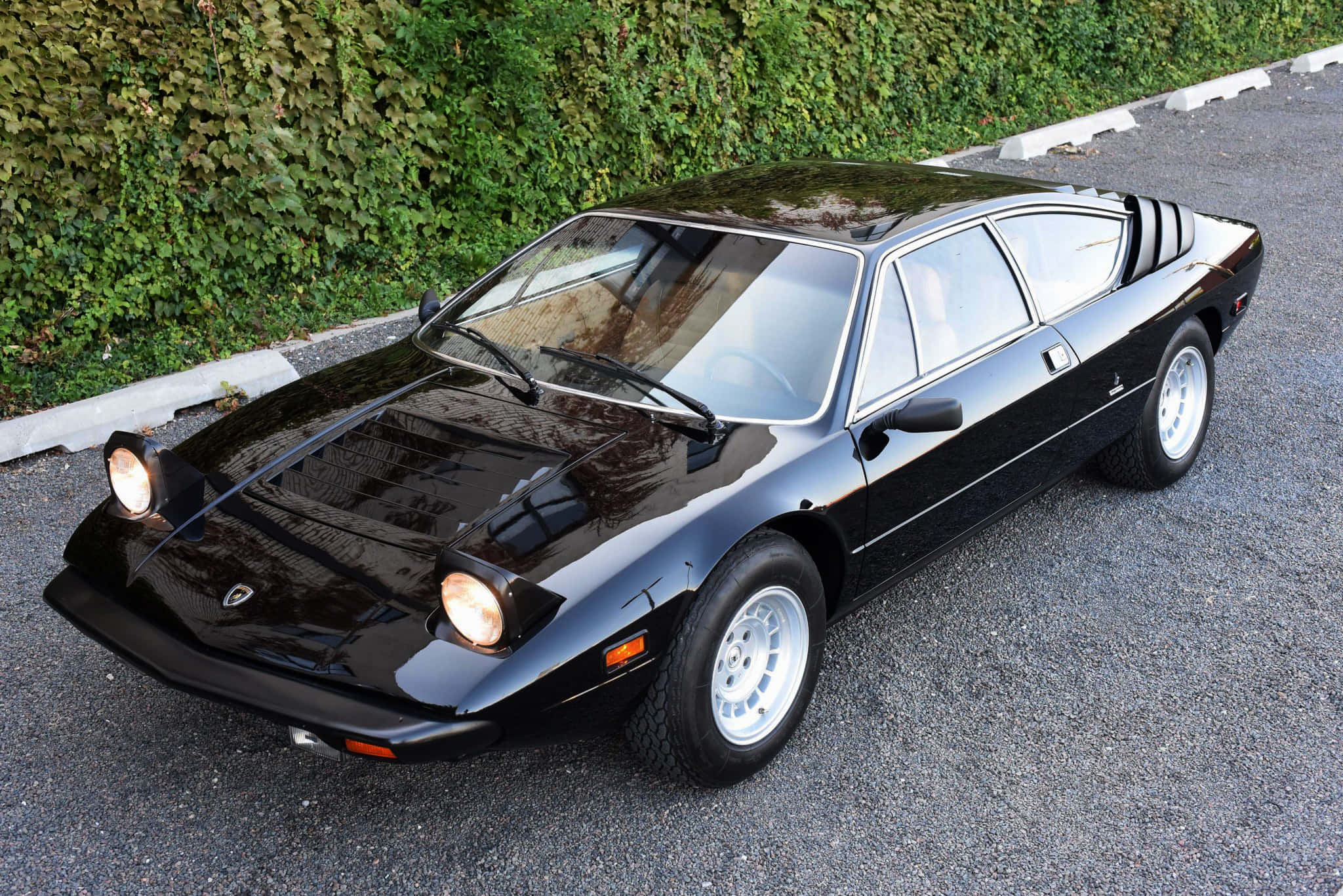 Lamborghini Urraco 桌布 壁紙 — 一輛時尚的黑色經典跑車，配白色車輪，停在碎石路上，旁邊是茂密的綠色樹籬。