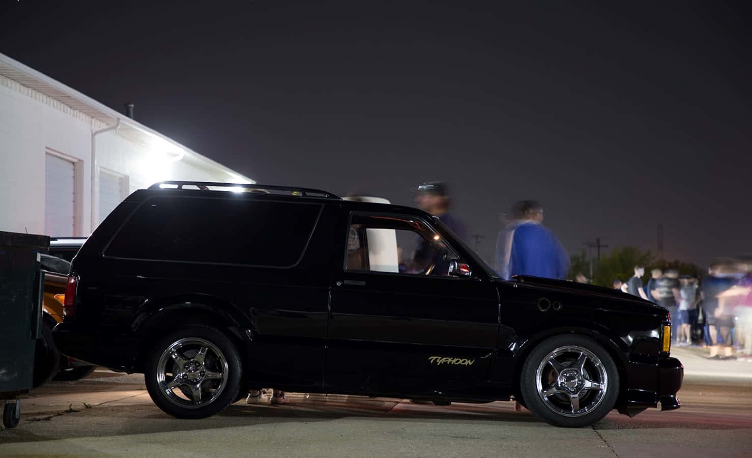 Gmc Typhoon 桌布 壁紙 — 一輛時尚的黑色定制SUV，配備鍍鉻輪圈，夜間在人工燈光下停車，附近有模糊的人群。