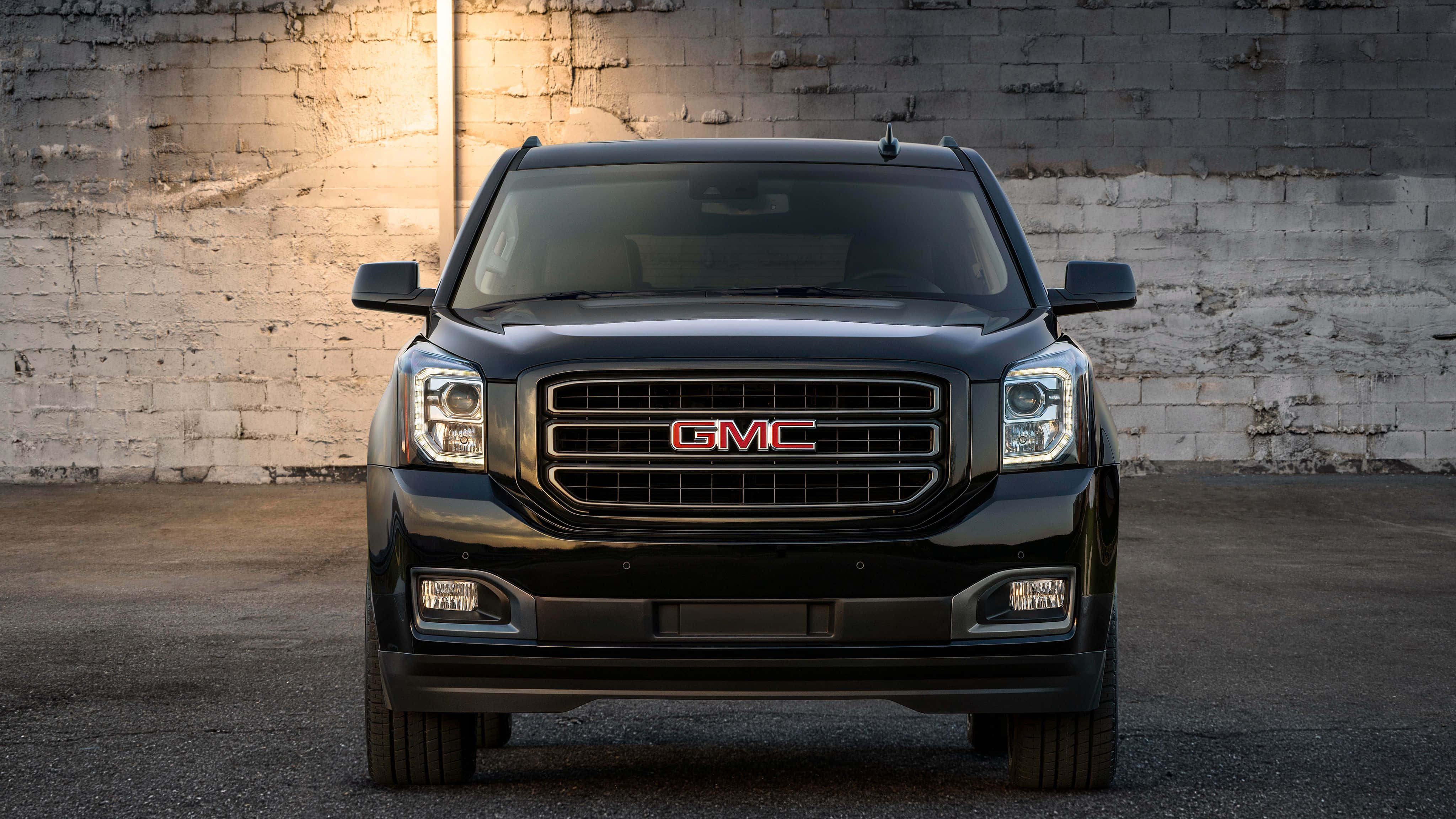 Gmc Yukon 桌布 壁紙 — 一輛時尚的黑色GMC SUV在溫暖的聚光燈下向前，背景是風化的白色磚牆。