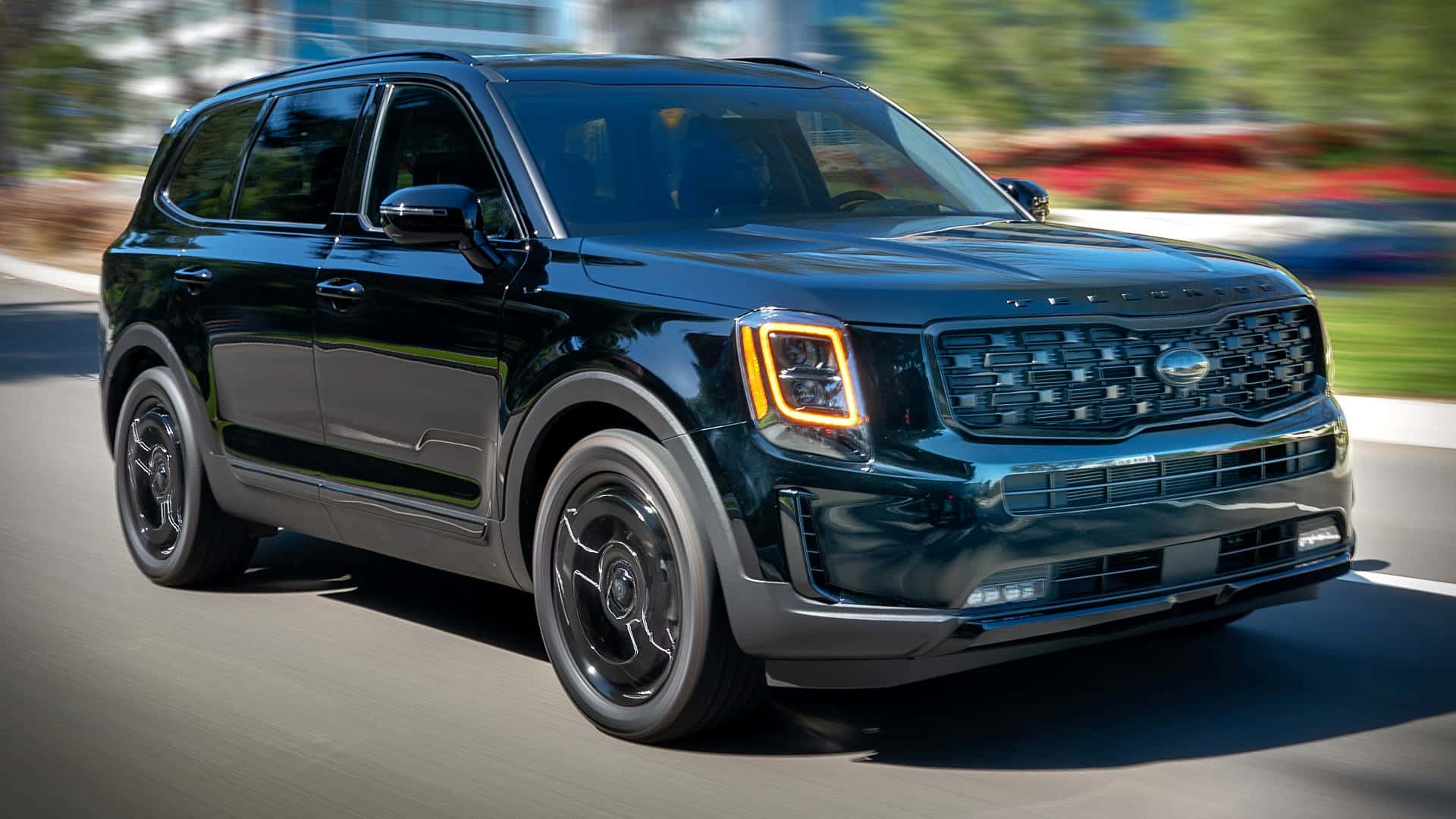 Kia Telluride 桌布 壁紙 — 一輛時尚的黑色Kia Telluride SUV在陽光明媚的道路上行駛，背景是模糊的綠色植物和色彩繽紛的花朵。