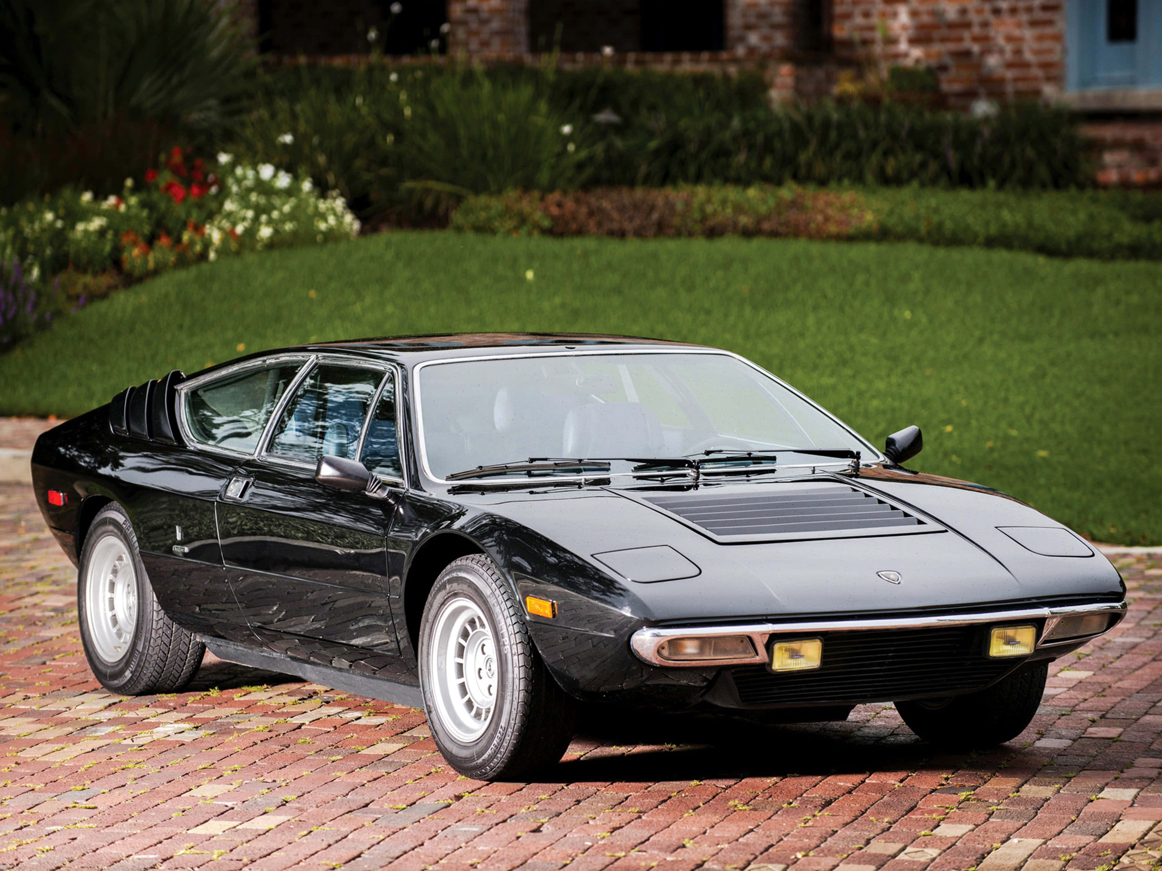 Lamborghini Urraco 桌布 壁紙 — 一架時尚的黑色藍寶堅尼Countach停放在紅磚車道上，背景是修剪整齊的草坪和磚屋。
