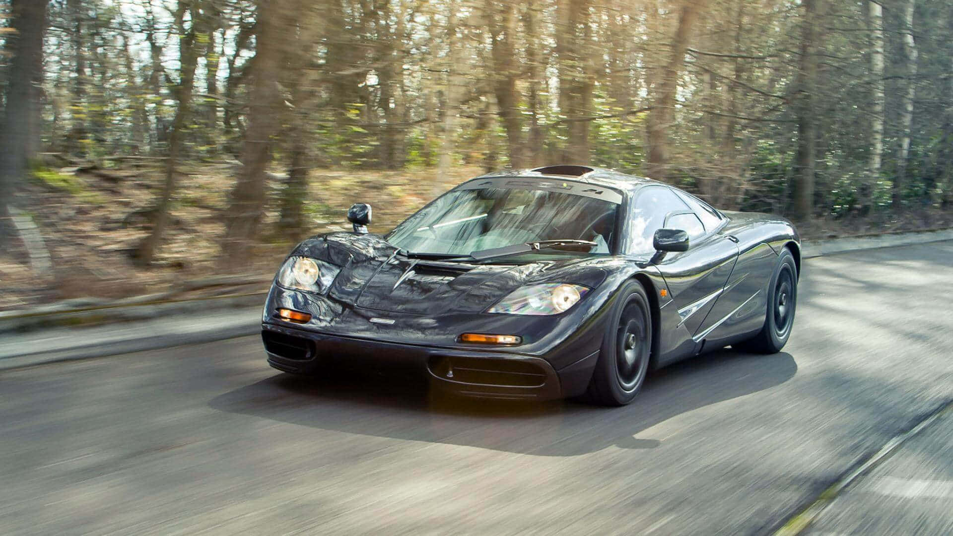 Mclaren F1 桌布 壁紙 — 一輛時尚的黑色McLaren F1在陽光明媚的森林道路上飛馳，樹木和瀝青上帶有動態模糊效果。