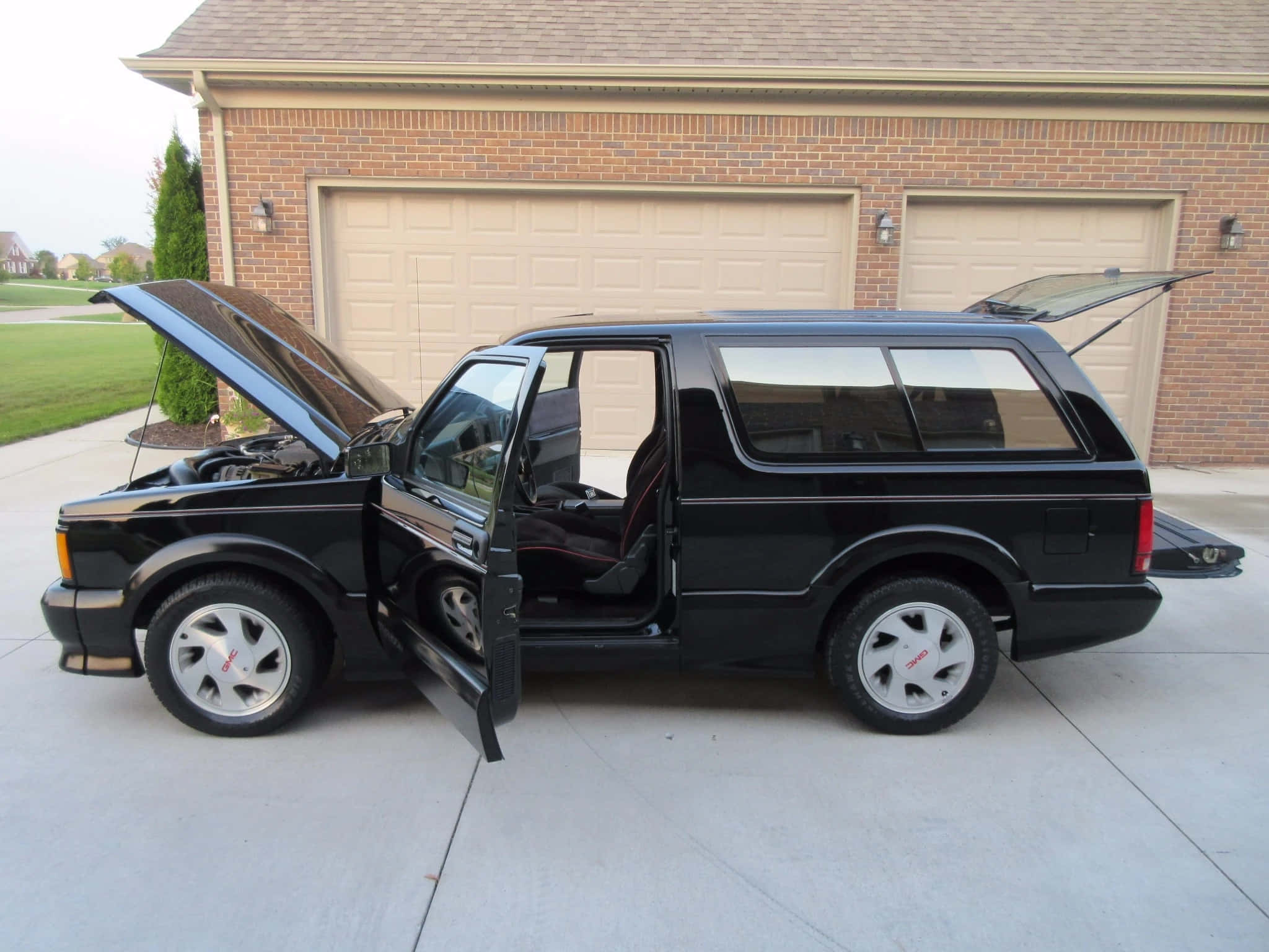 Gmc Typhoon 桌布 壁紙 — 一輛時尚的黑色SUV，車門和引擎蓋打開，停在磚房前的混凝土車道上。