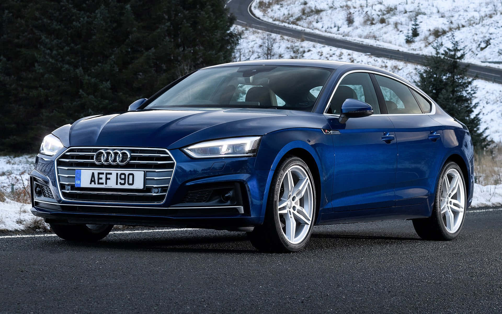 Audi A5 桌布 壁紙 — 一輛時尚的藍色奧迪轎車在明亮的日光下，在蜿蜒的雪路上閃閃發光，兩旁是常青樹。