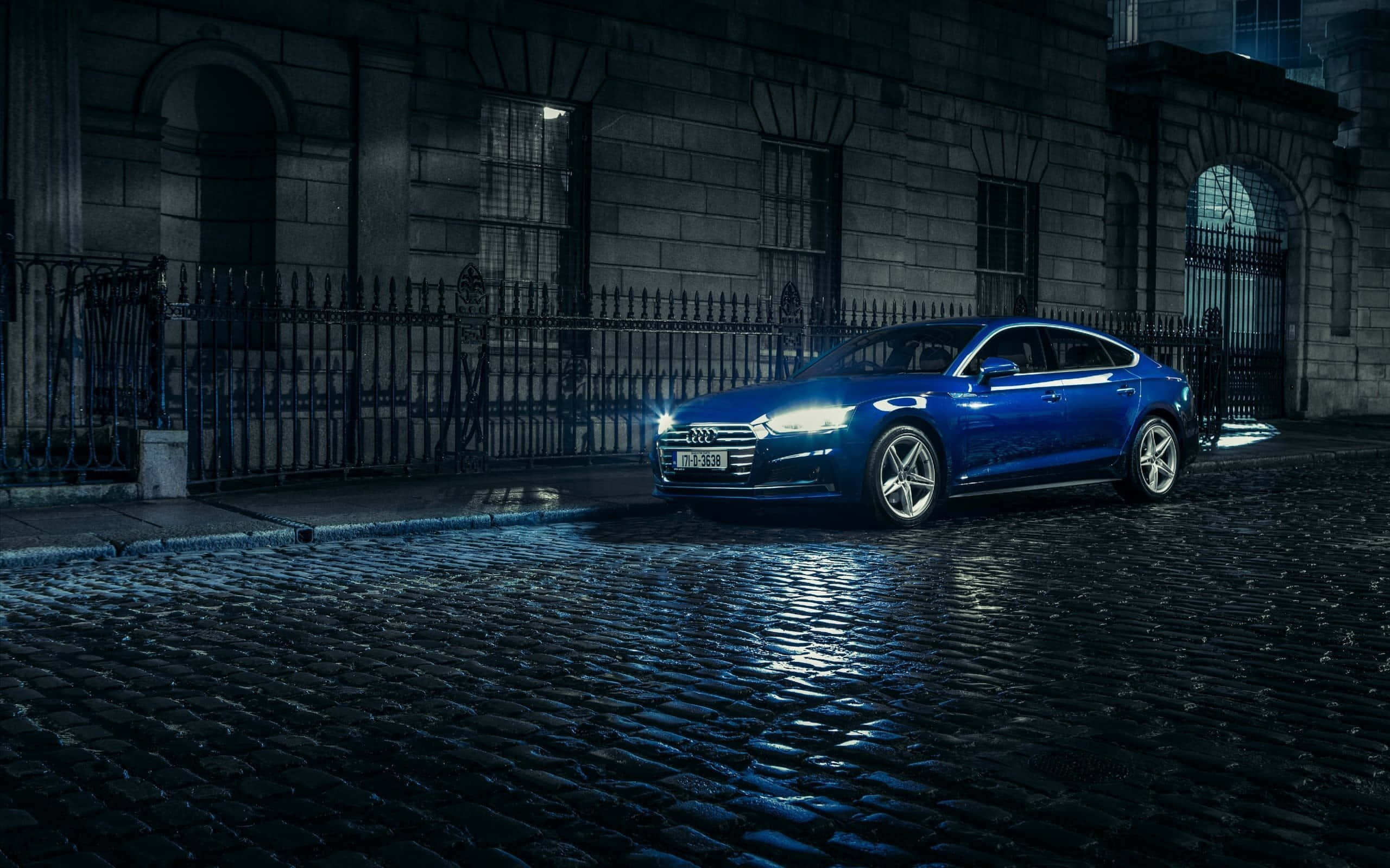 Audi A5 桌布 壁紙 — 一輛時尚的藍色奧迪轎車在夜間濕滑的鵝卵石街道上，在石頭建築旁，於車頭燈下閃耀。
