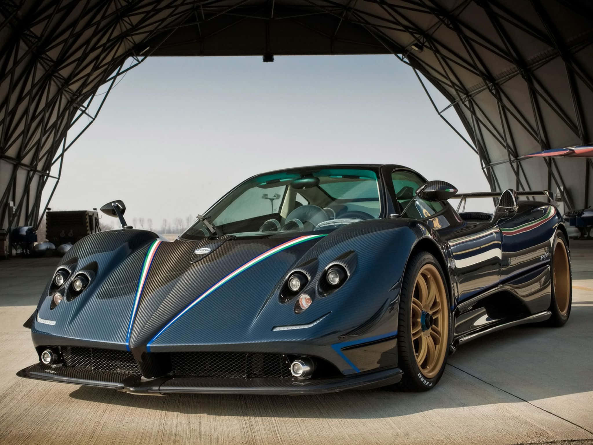 Pagani Zonda Tricolore 桌布 壁紙 — 一輛時尚的藍色碳纖維帕加尼跑車，配備金色輪圈，停放在現代金屬機庫下。