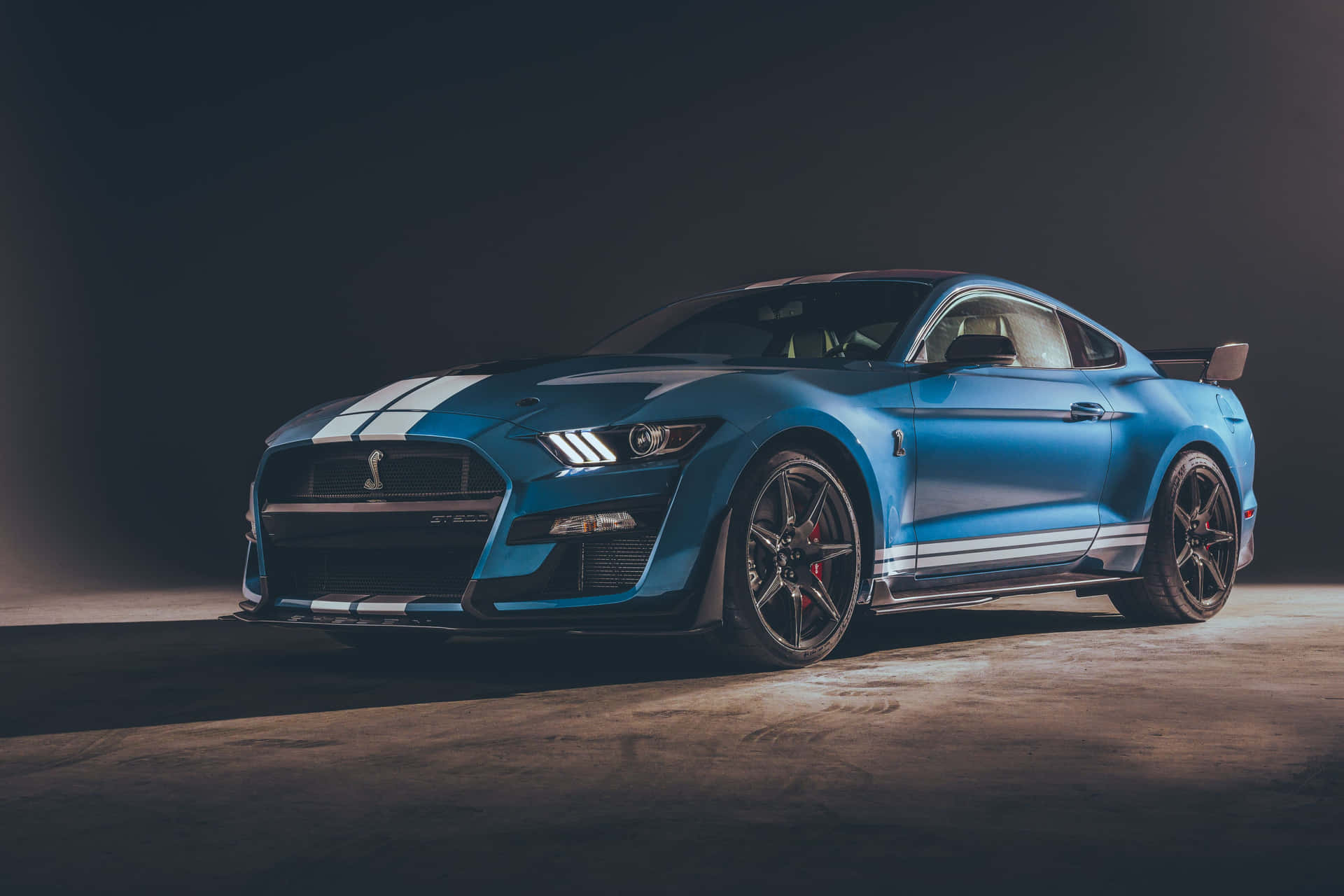  壁紙 — 一輛時尚的藍色Shelby GT500跑車帶有白色賽車條紋，在黑暗的攝影棚背景下戲劇性地照亮。