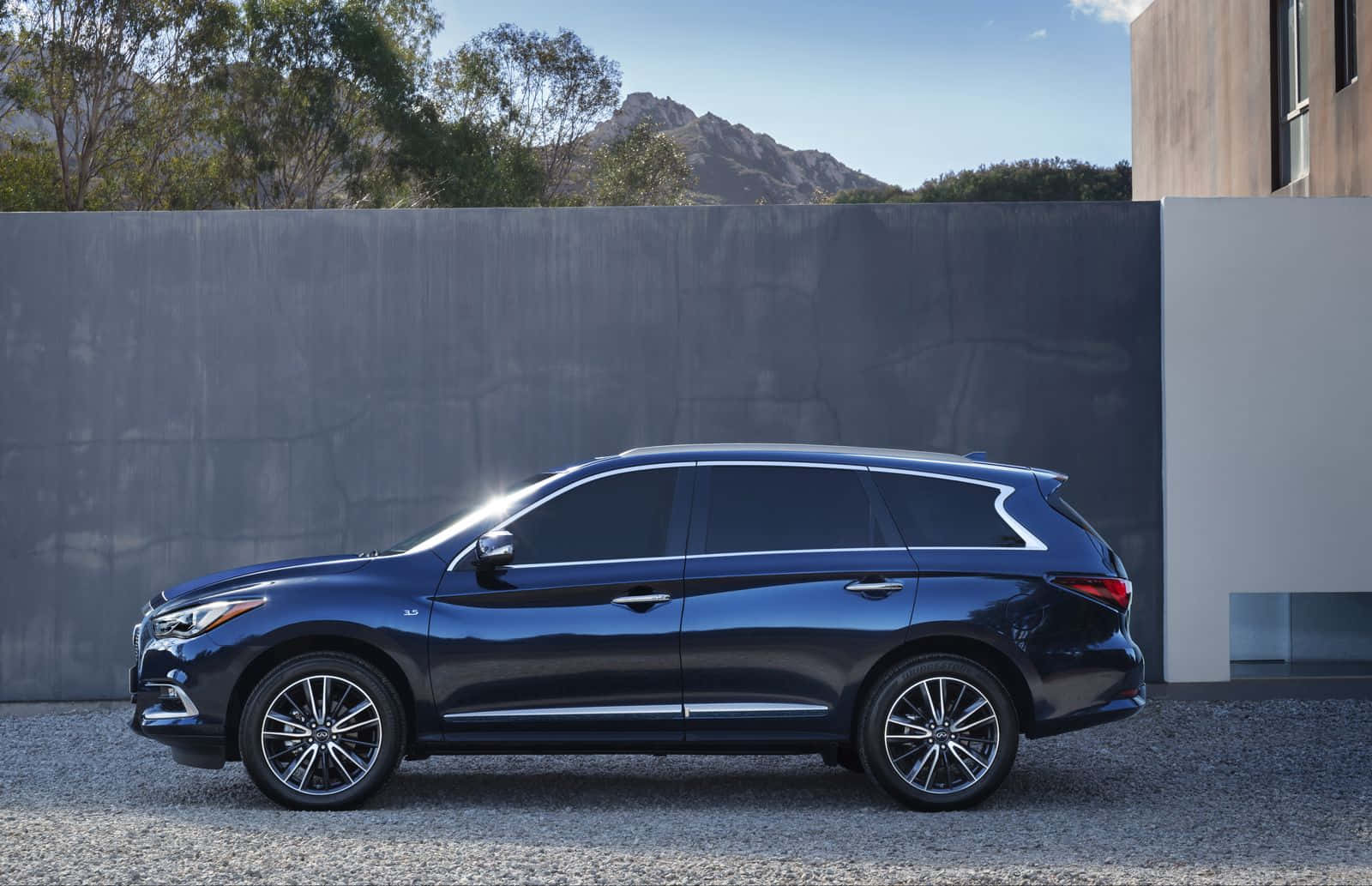 Infiniti Qx60 桌布 壁紙 — 一輛時尚深藍色SUV停在碎石上，旁邊是現代混凝土牆，背景有山。