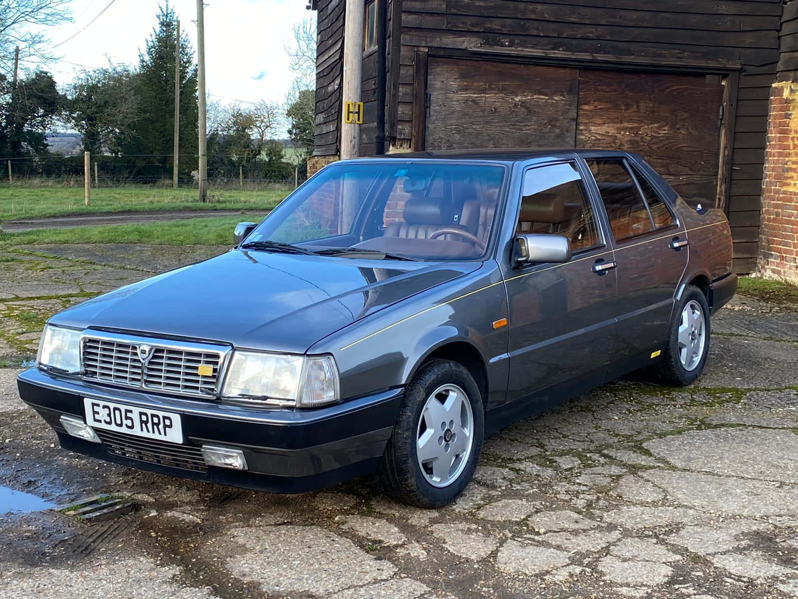  壁紙 — 一輛時尚深灰色 Lancia Thema 轎車停放在裂開的石頭車道上，旁邊是質樸的木車庫。