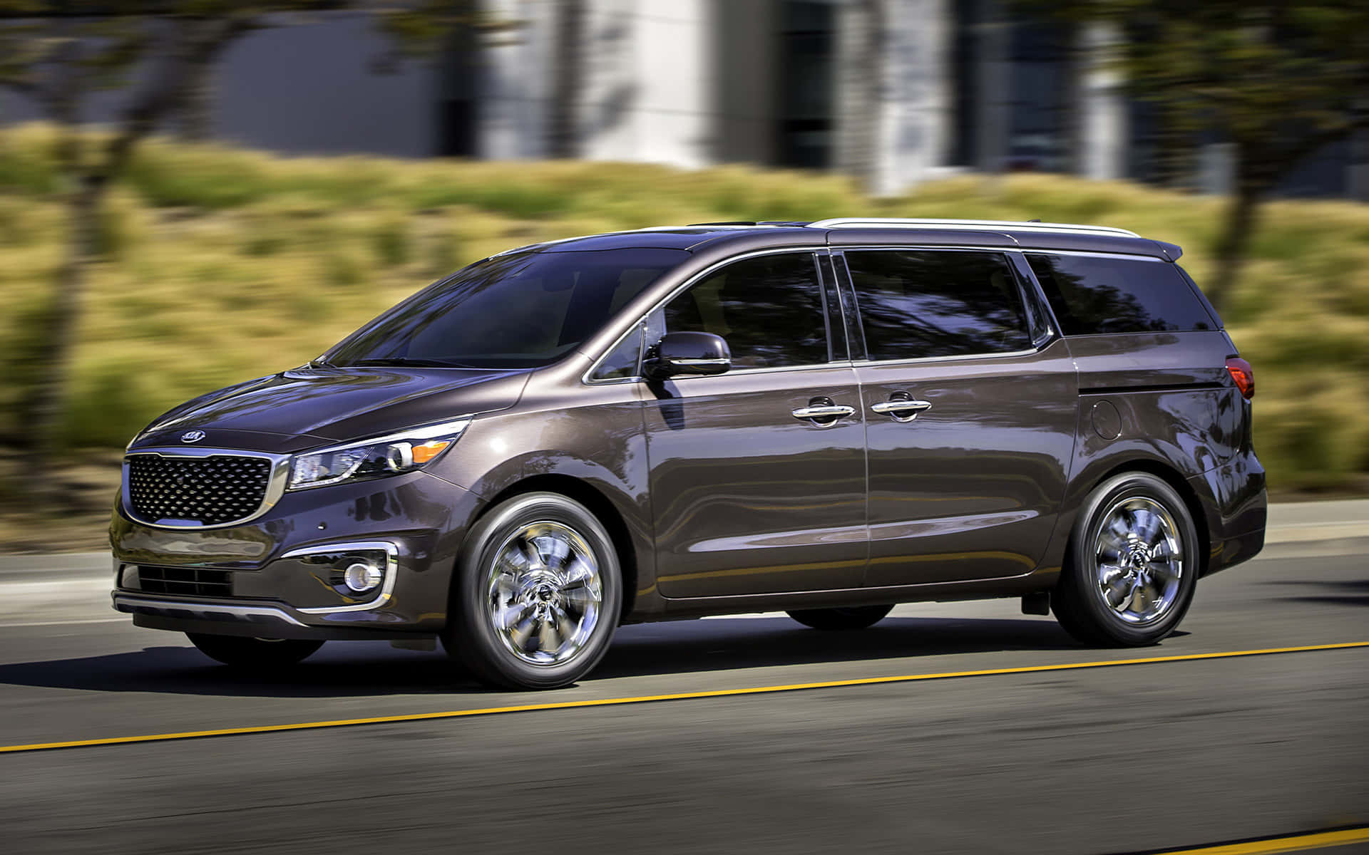 Kia Sedona 桌布 壁紙 — 一輛時尚的深灰色小型貨車在陽光明媚的道路上行駛，背景有模糊的綠化和建築物。