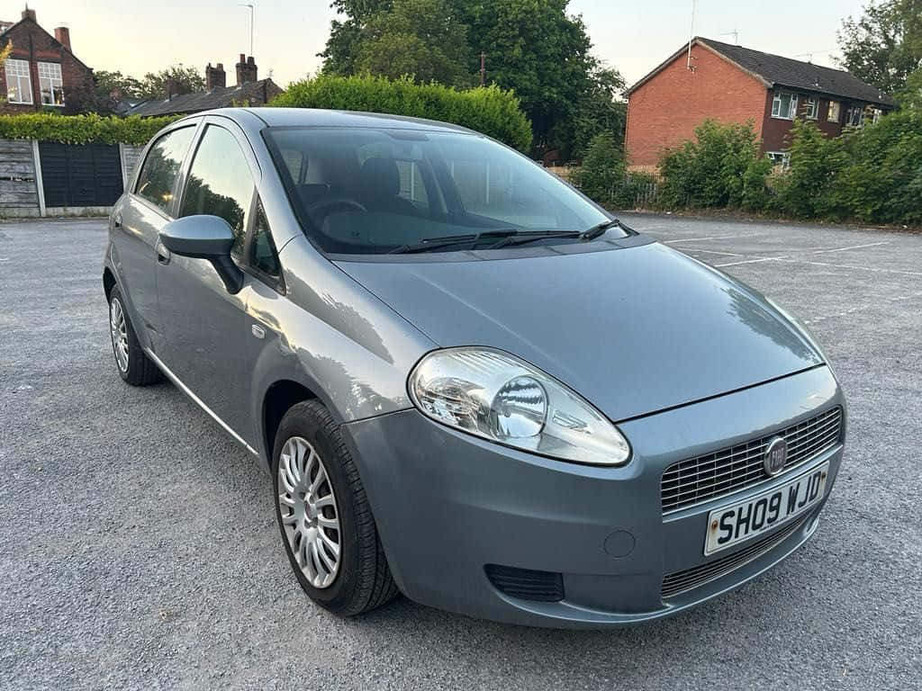 Fiat Punto 桌布 壁紙 — 一輛時尚的灰色 Fiat Punto 停放在碎石地上，背景是黃昏時分的磚房和綠色籬笆。