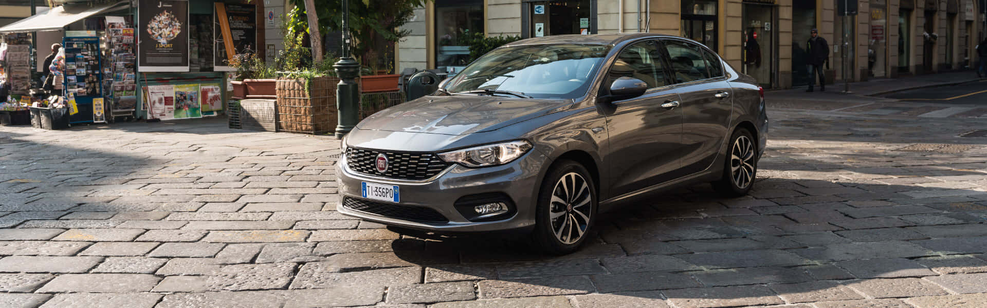 Fiat Tipo 桌布 壁紙 — 一架流線型灰色飛雅特房車停泊在歐洲小鎮陽光普照的鵝卵石街道上，附近有商店和行人。