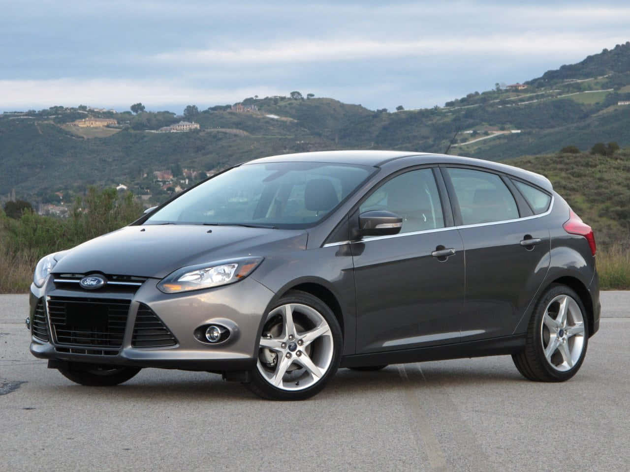 Ford Focus 桌布 壁紙 — 一輛時尚的灰色福特福克斯掀背車停在鋪砌的道路上，背景是起伏的綠色山丘和散落的房屋，天空多雲。