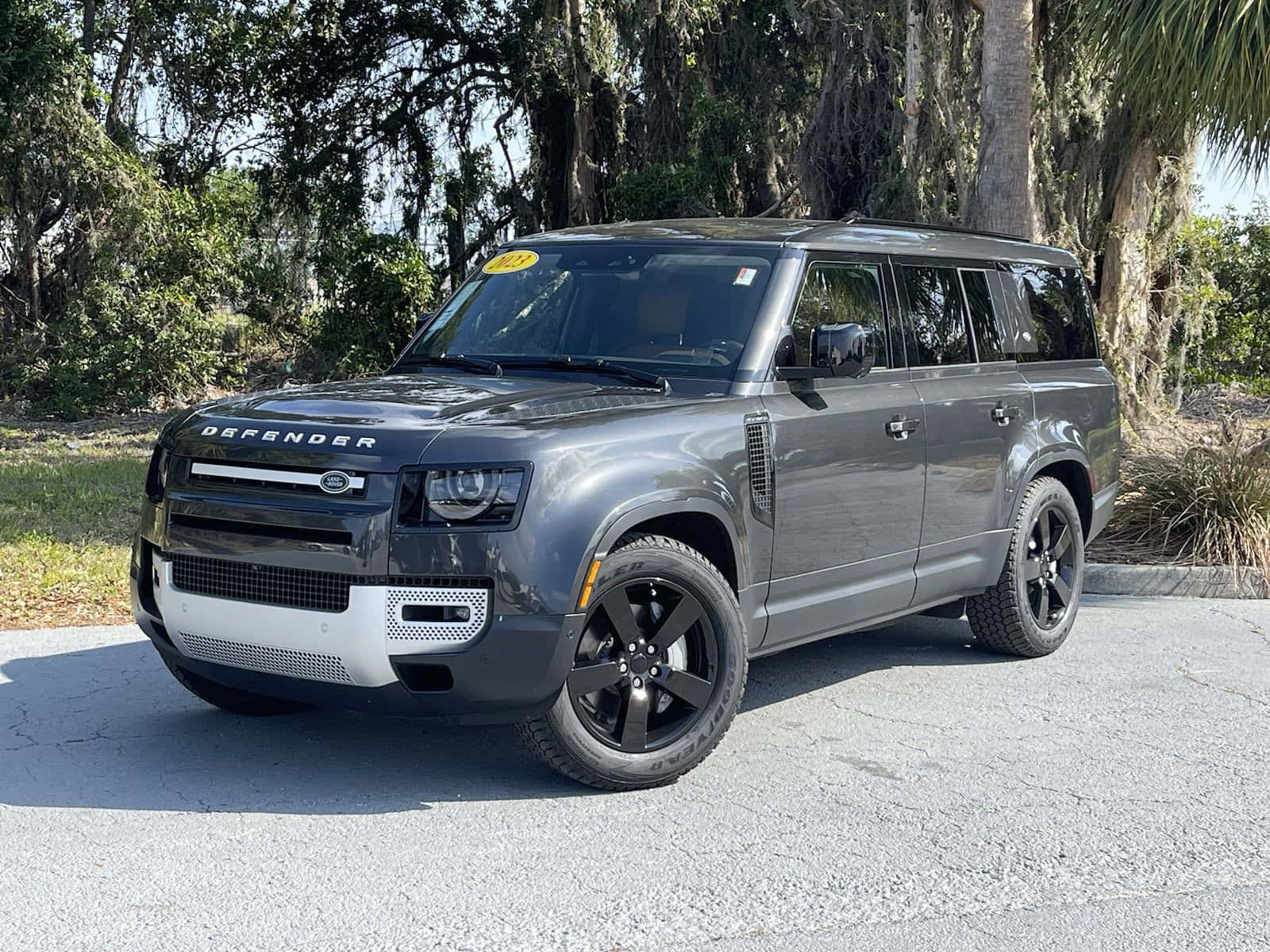 Land Rover Defender 桌布 壁紙 — 一輛時尚的灰色 Land Rover Defender SUV 停放在裂開的路面上，背景是明亮的日光和茂密的綠樹。