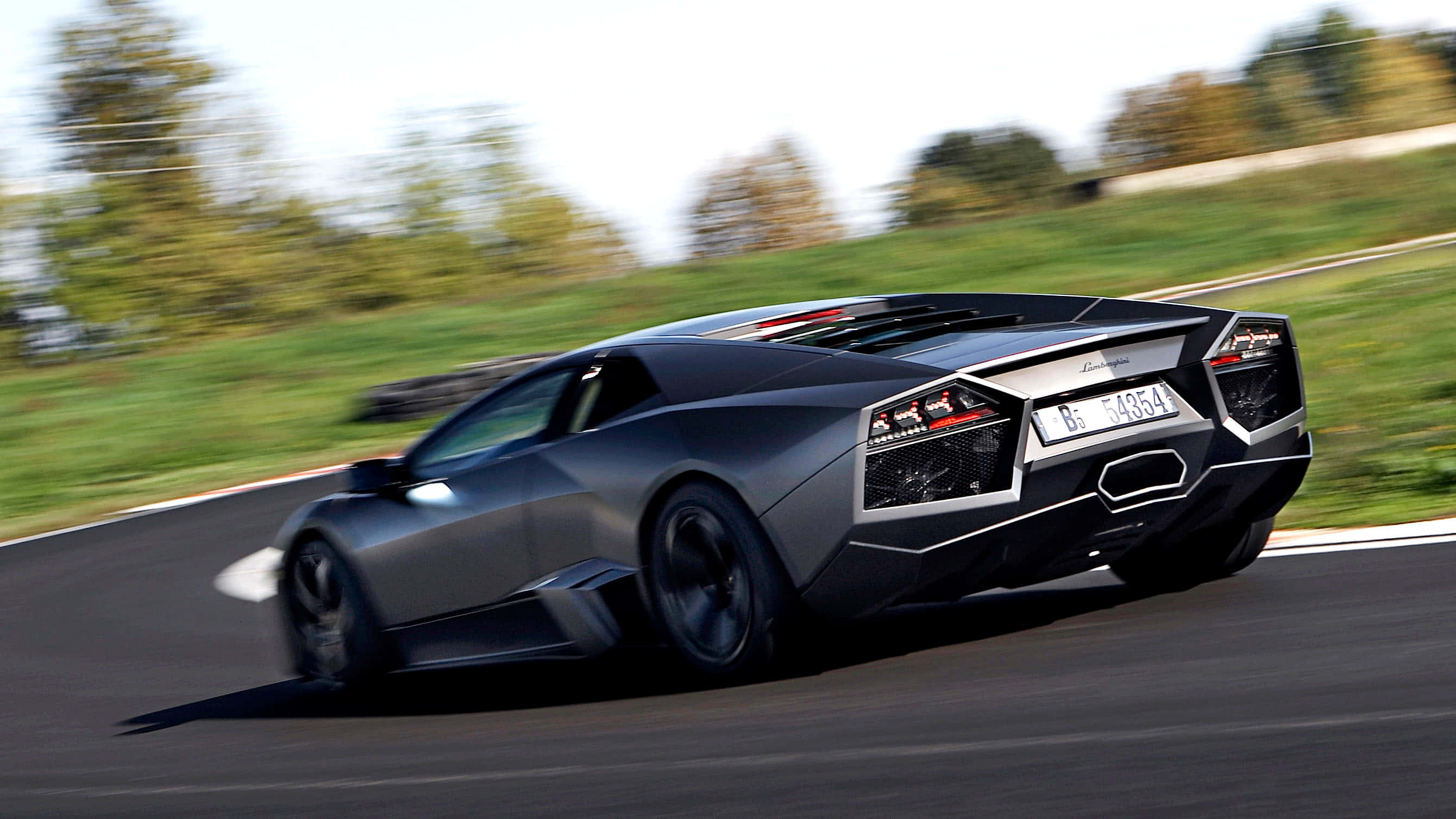 Lamborghini Reventón 桌布 壁紙 — 一輛時尚的啞光灰色藍寶堅尼Reventon沿著彎曲的瀝青道路疾馳，背景有模糊的綠色樹木。