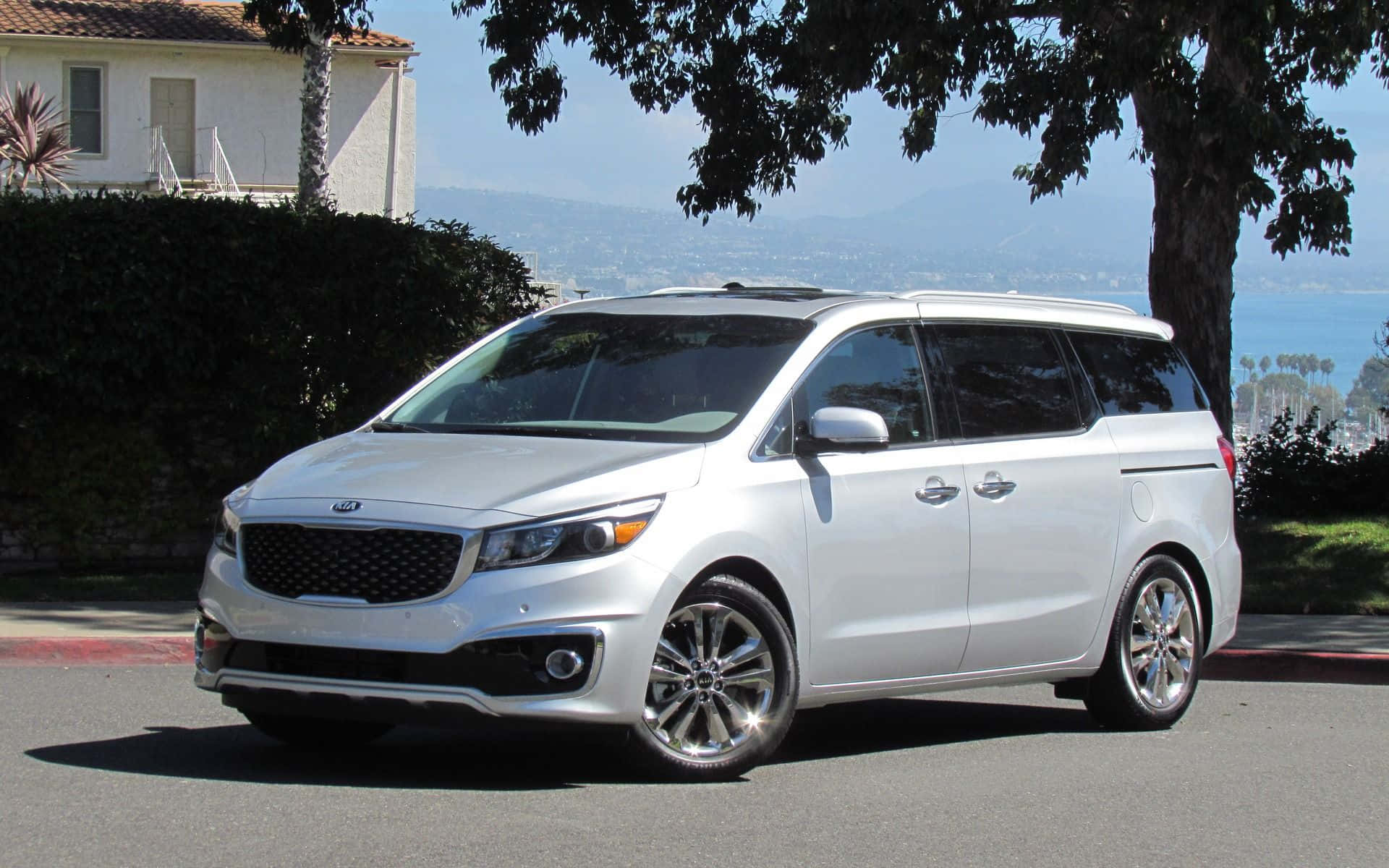 Kia Sedona 桌布 壁紙 — 一架時尚的銀色Kia小型貨車停放在陽光明媚的街道上，背景有樹木、遠山和海洋。