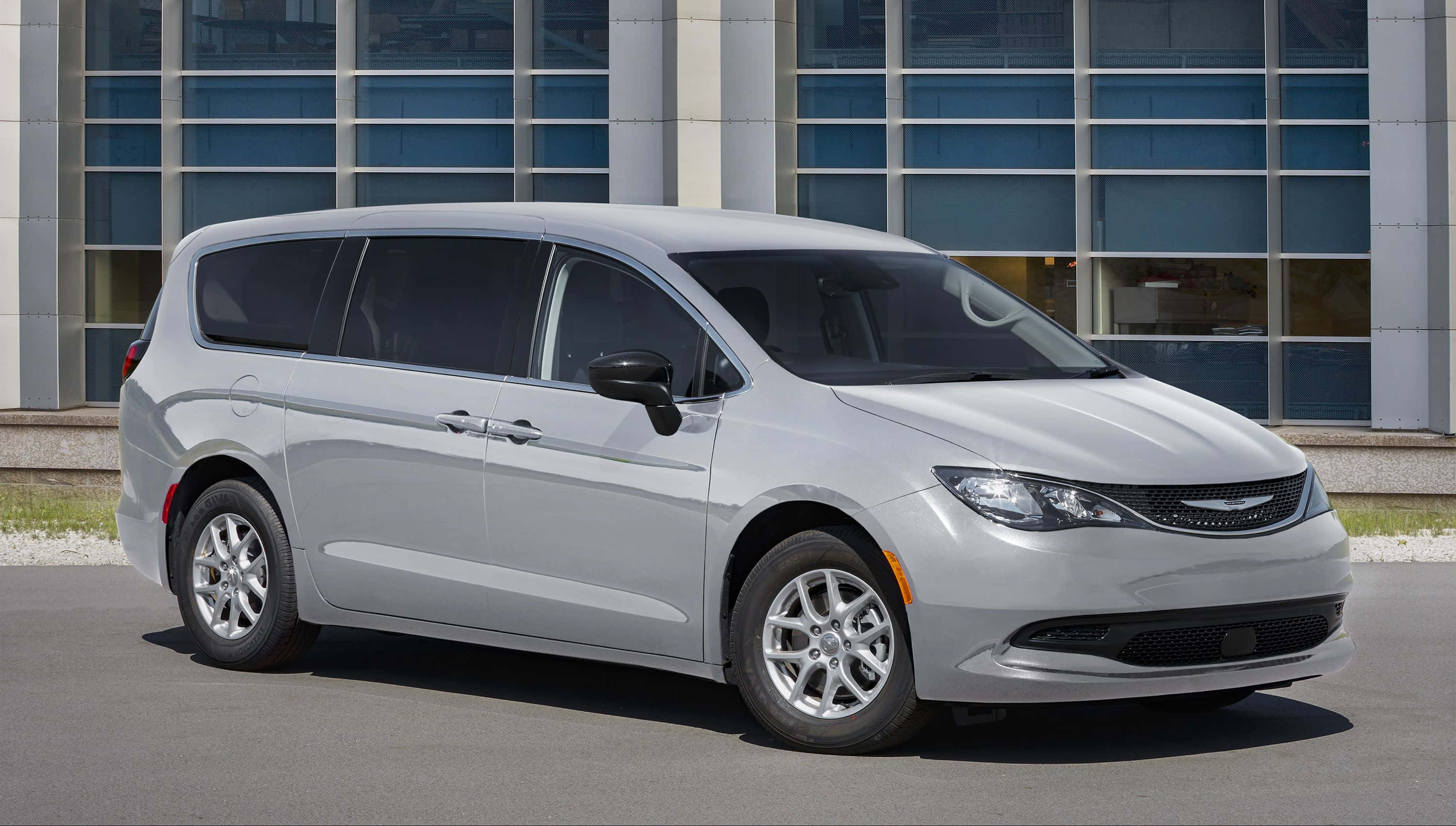 Chrysler Voyager 桌布 壁紙 — 一輛時尚的銀色小型貨車停在現代玻璃建築前，陽光明媚。