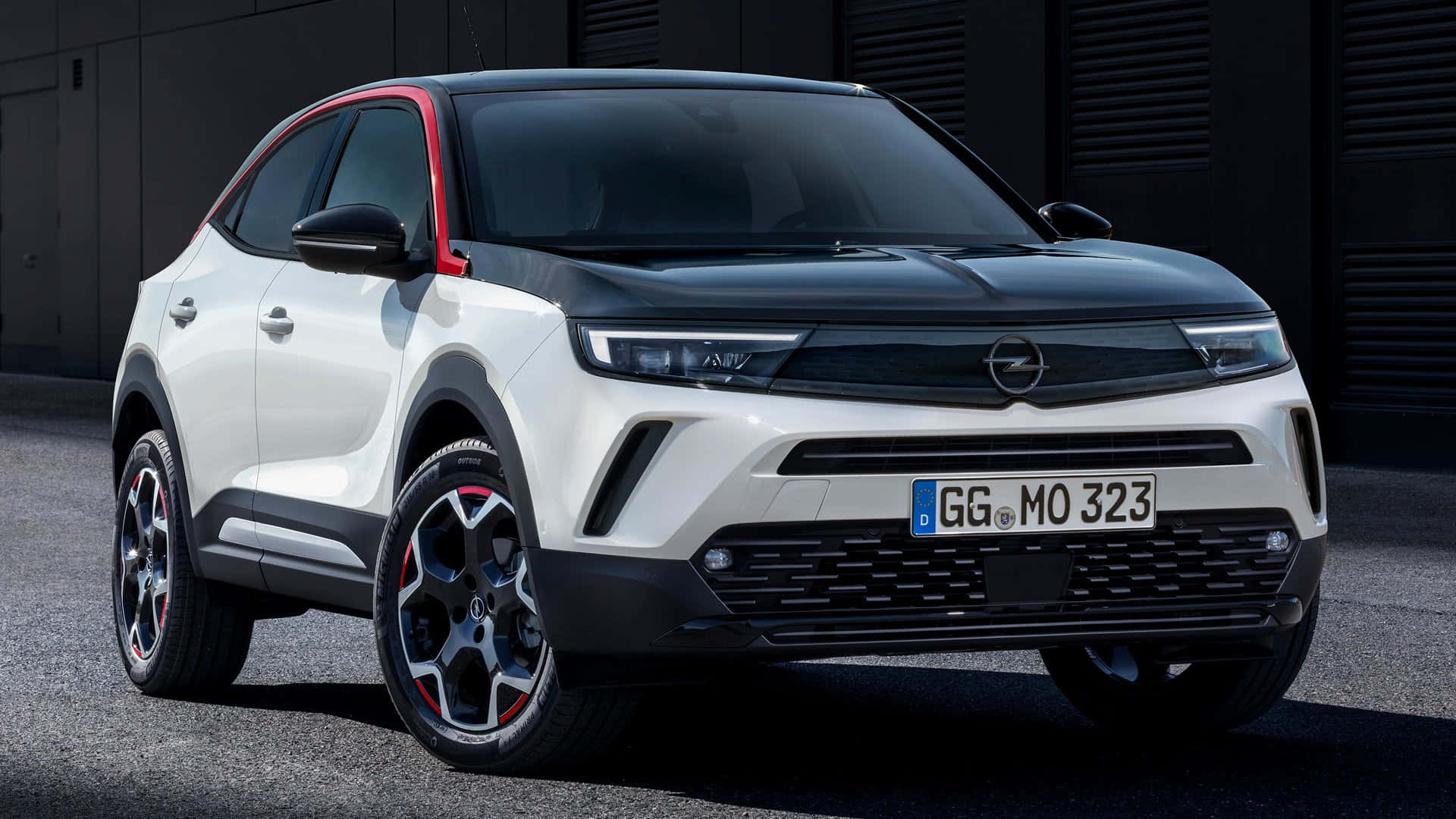 Opel Mokka 桌布 壁紙 — 一輛時尚的白色和黑色歐寶 Mokka X SUV，帶有紅色點綴，停在瀝青上，背景是深色工業牆。