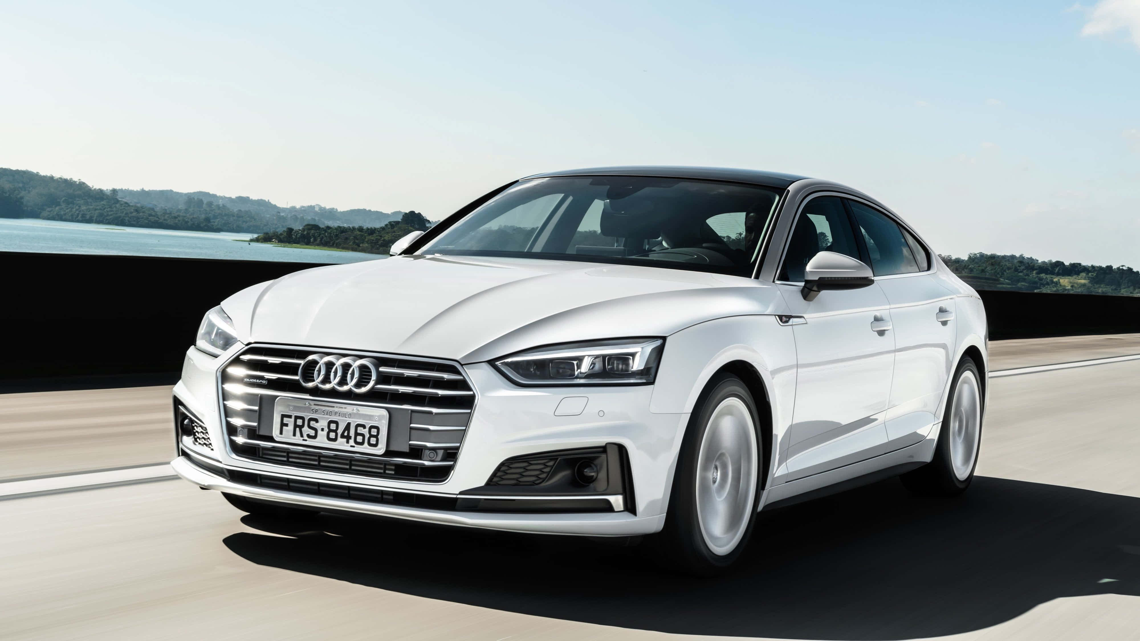 Audi A5 桌布 壁紙 — 一輛時尚的白色奧迪轎車在高速公路上行駛，背景是風景如畫的湖泊和森林覆蓋的山丘，天空清澈湛藍。