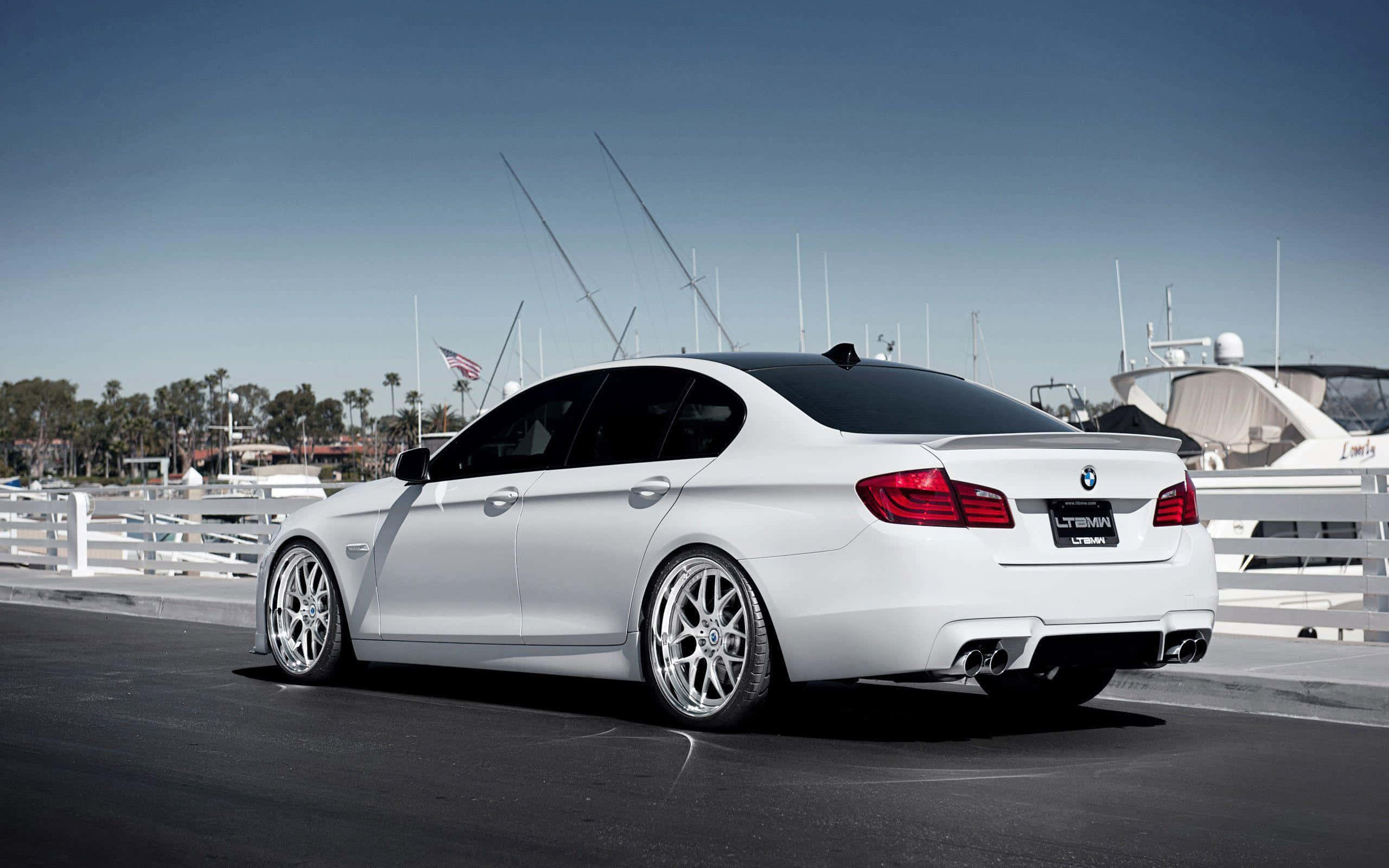 Bmw M5 桌布 壁紙 — 一輛時尚的白色BMW轎車配備定制輪圈，停泊在碼頭旁，旁邊是豪華遊艇，天空清澈湛藍。