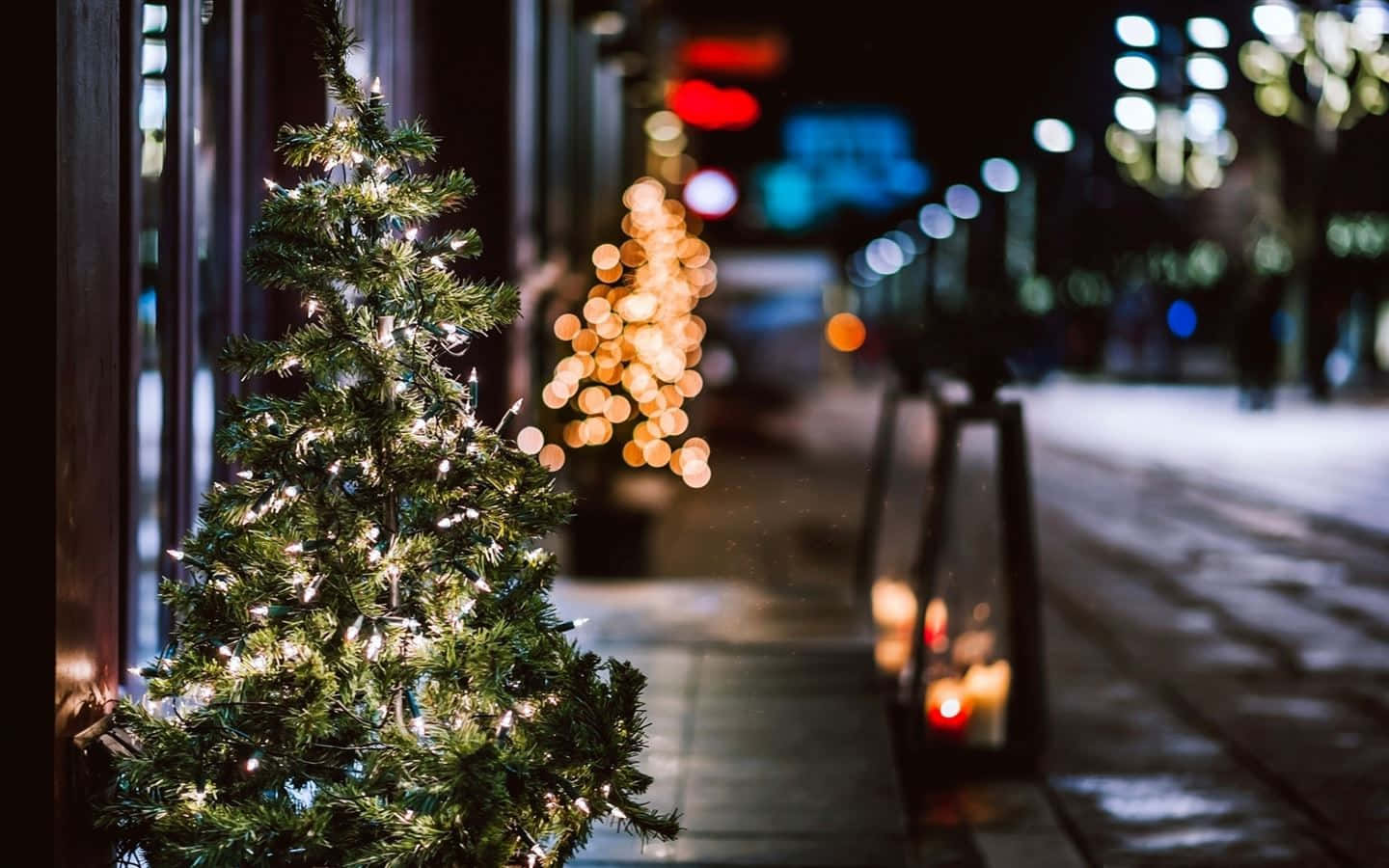  壁紙 — 一棵小裝飾聖誕樹在雪夜的城市人行道上閃爍著燈串，背景是散景的路燈。