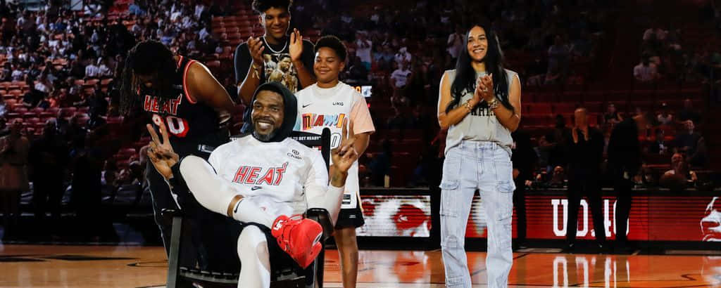  壁紙 — 一位微笑的籃球運動員身穿白色Heat球衣坐在球場中央做出和平手勢，家人和隊友在他身後鼓掌。
