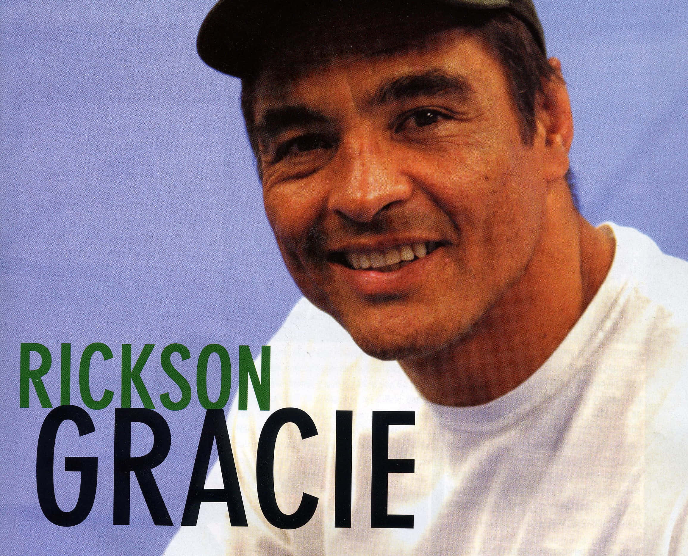 Rickson Gracie 桌布 壁紙 — 一個戴帽子和白色T恤的微笑男子，背景為純藍色，他的名字以粗體綠色和黑色文字顯示。