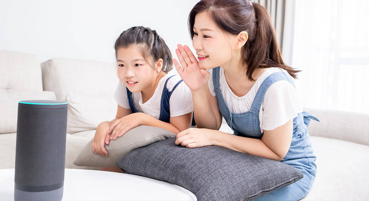 穿著連身褲的母親和女兒在沙發上向智慧音箱低語。 — Google 壁紙