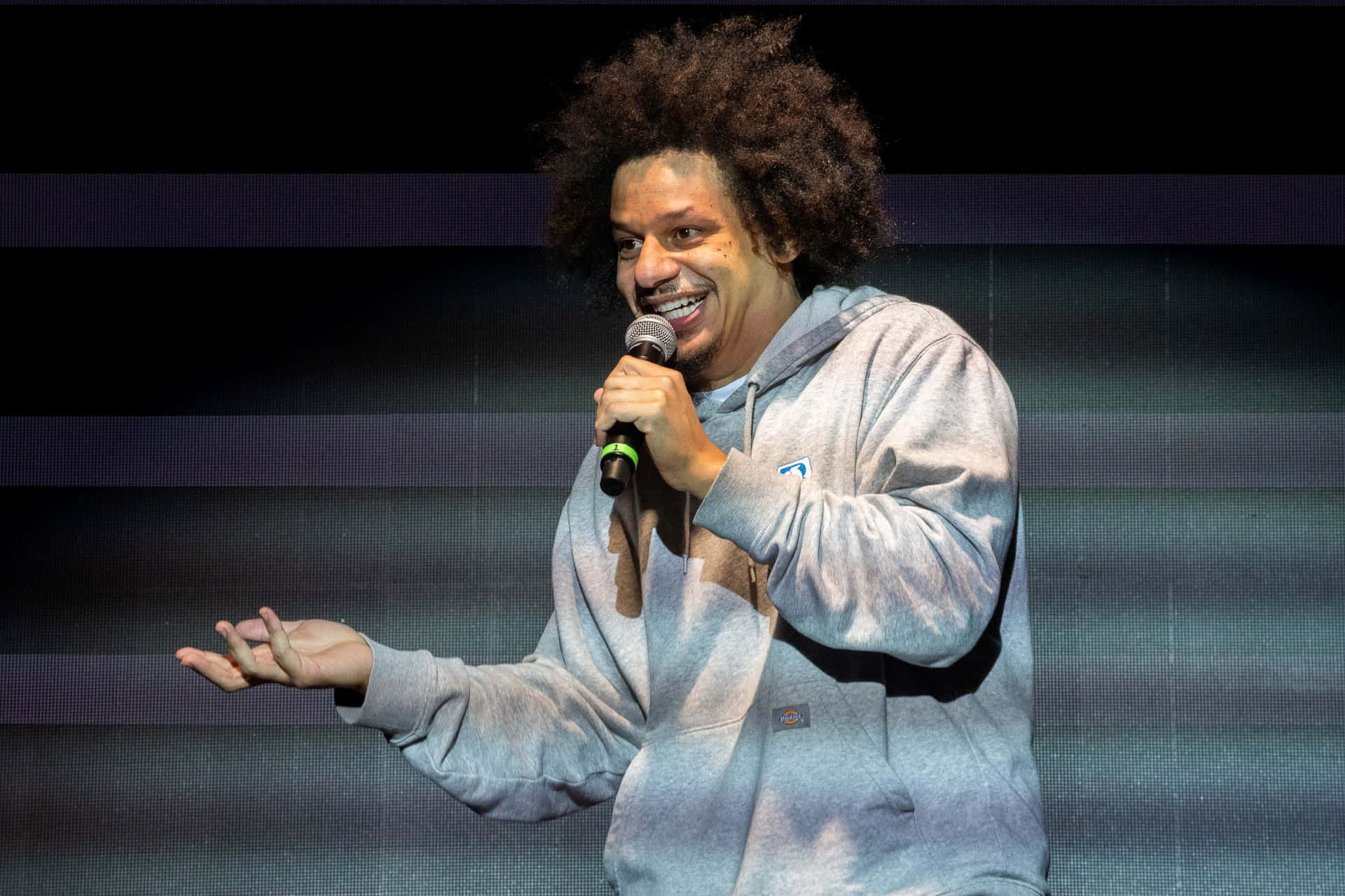  壁紙 — 一位笑容滿面、留著捲髮的表演者在舞台上，背後是深色條紋背景，手持麥克風。