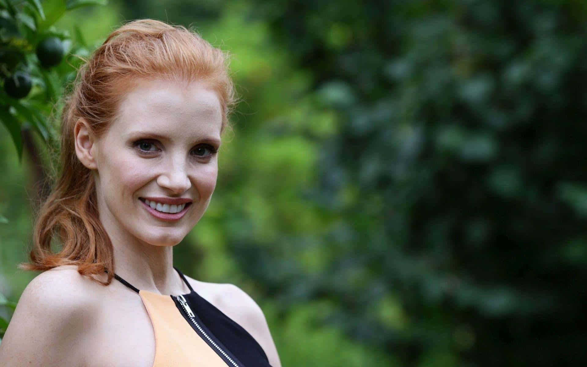 jessica chastain 桌布 壁紙 — 一位微笑的紅髮女性穿著黑色和橙色上衣，站在茂密的綠色樹葉背景前。