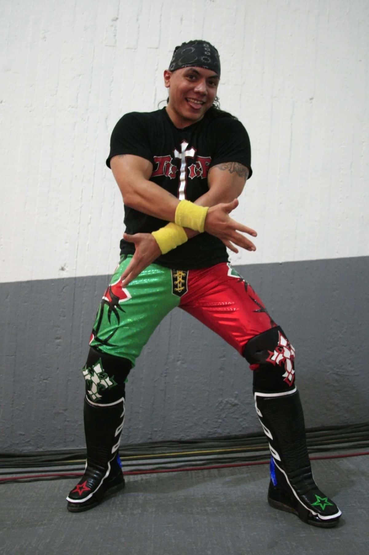  壁紙 — 一位微笑的摔角手穿著色彩繽紛的紅、綠、黑色服裝，在素牆前擺出動感姿勢。