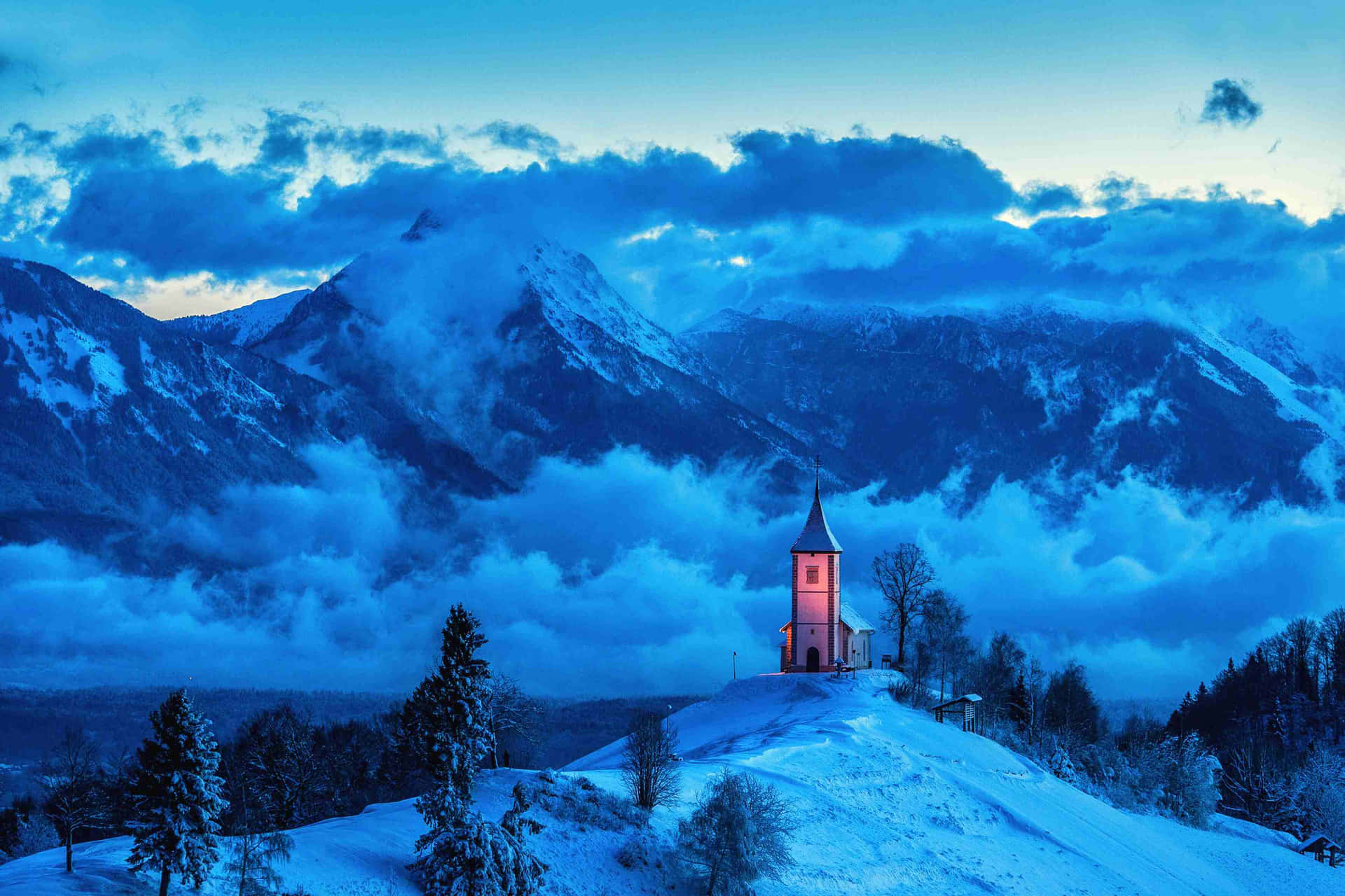  壁紙 — 一座被雪覆蓋的小教堂在山頂上溫暖地發光，背景是暮色中戲劇性的藍色山脈。