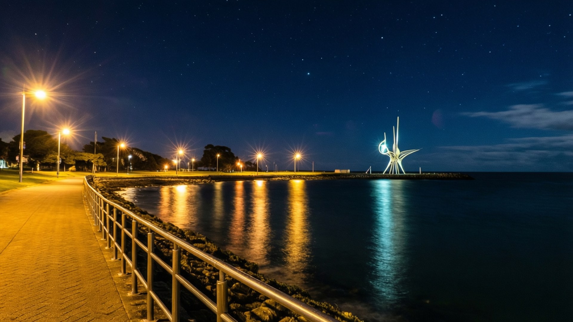 New Plymouth New Zealand 桌布 壁紙 — 星空下的夜晚海岸步道，溫暖的路燈照亮平靜的水面，倒映在發光的現代雕塑旁。