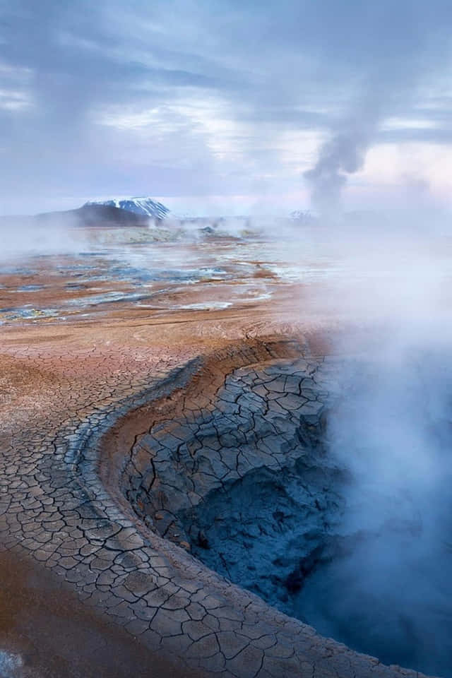  壁紙 — 一個冒著蒸汽的地熱火山口，龜裂的地面，薄霧在陰天中升起，遠處有積雪的山峰。
