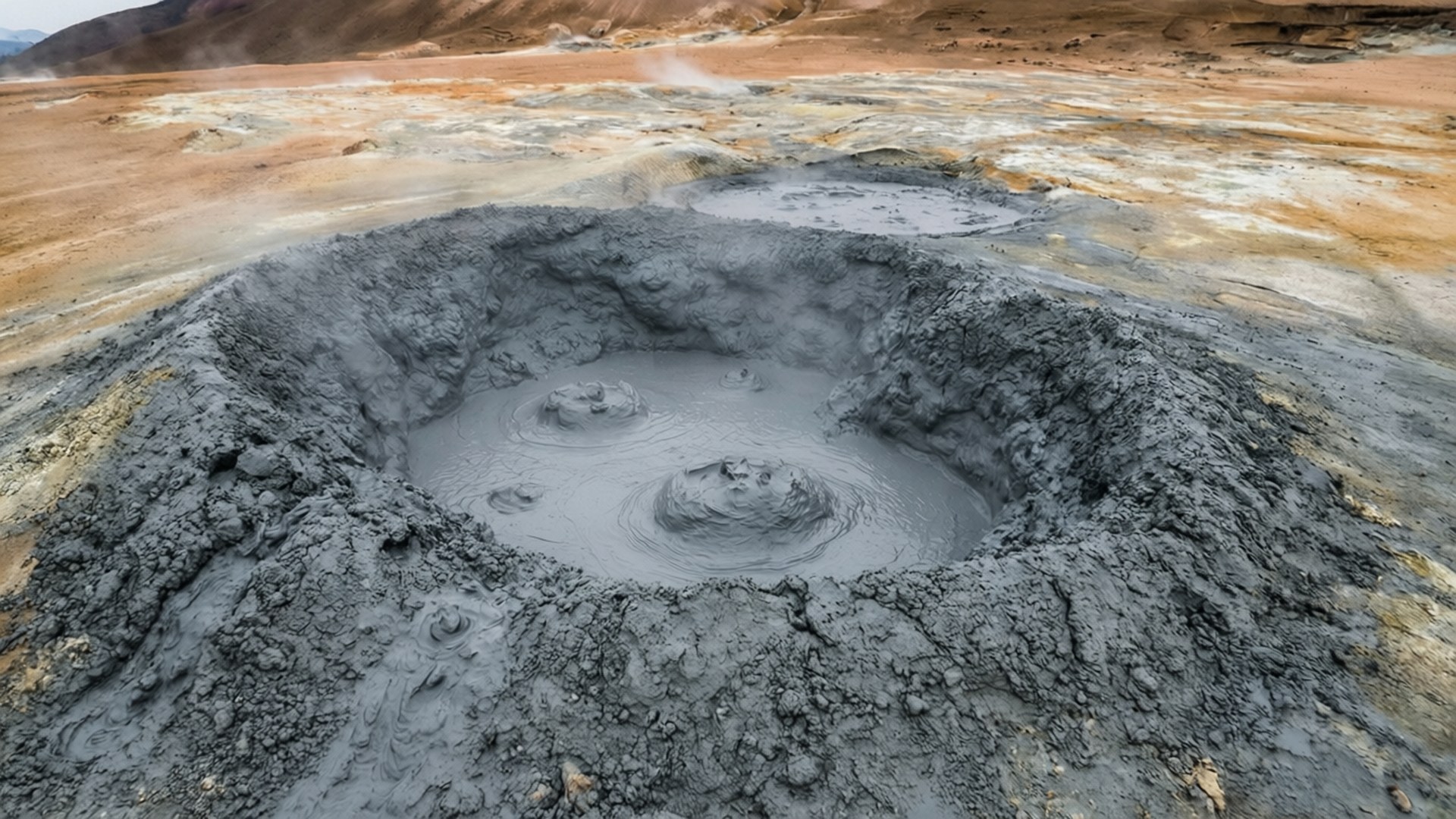 Buzau Muddy Volcanoes 壁紙 — 一個冒著蒸汽的灰色泥鍋在貧瘠的火山景觀中冒泡翻騰，帶有赭色和棕色的大地色調。