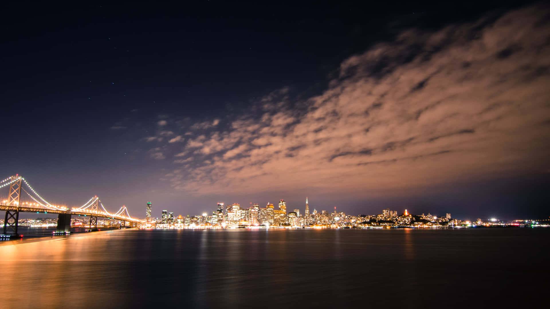  壁紙 — 令人驚嘆的城市天際線夜景，一座燈火通明的橋樑橫跨平靜的水面，上方是繁星點點的天空和發光的雲彩。