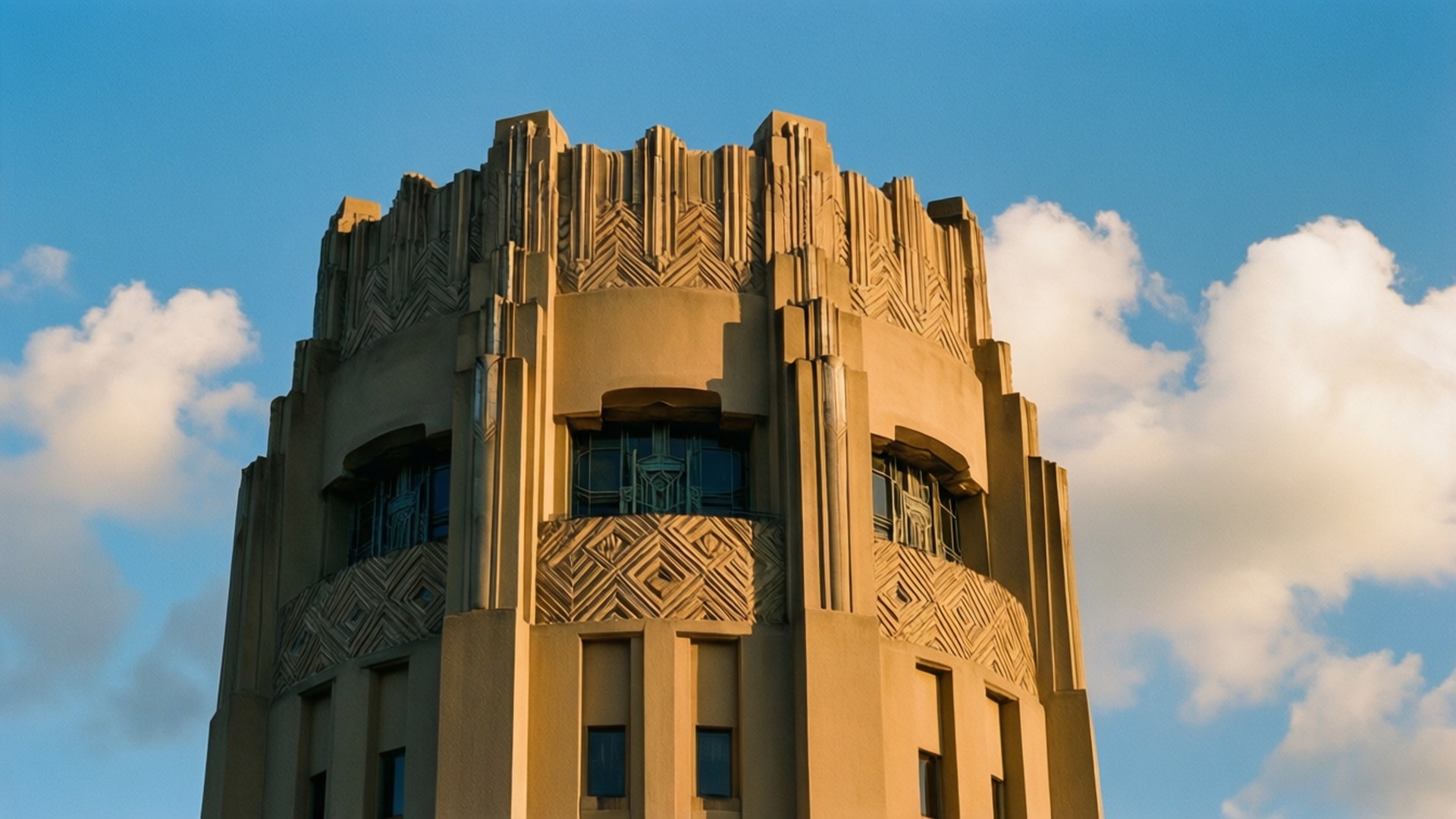 Bok Tower Art Deco 壁紙 — 一座陽光普照的裝飾藝術風格塔樓，帶有幾何雕刻和藍色窗戶，矗立在明亮的藍天和蓬鬆的白雲下。