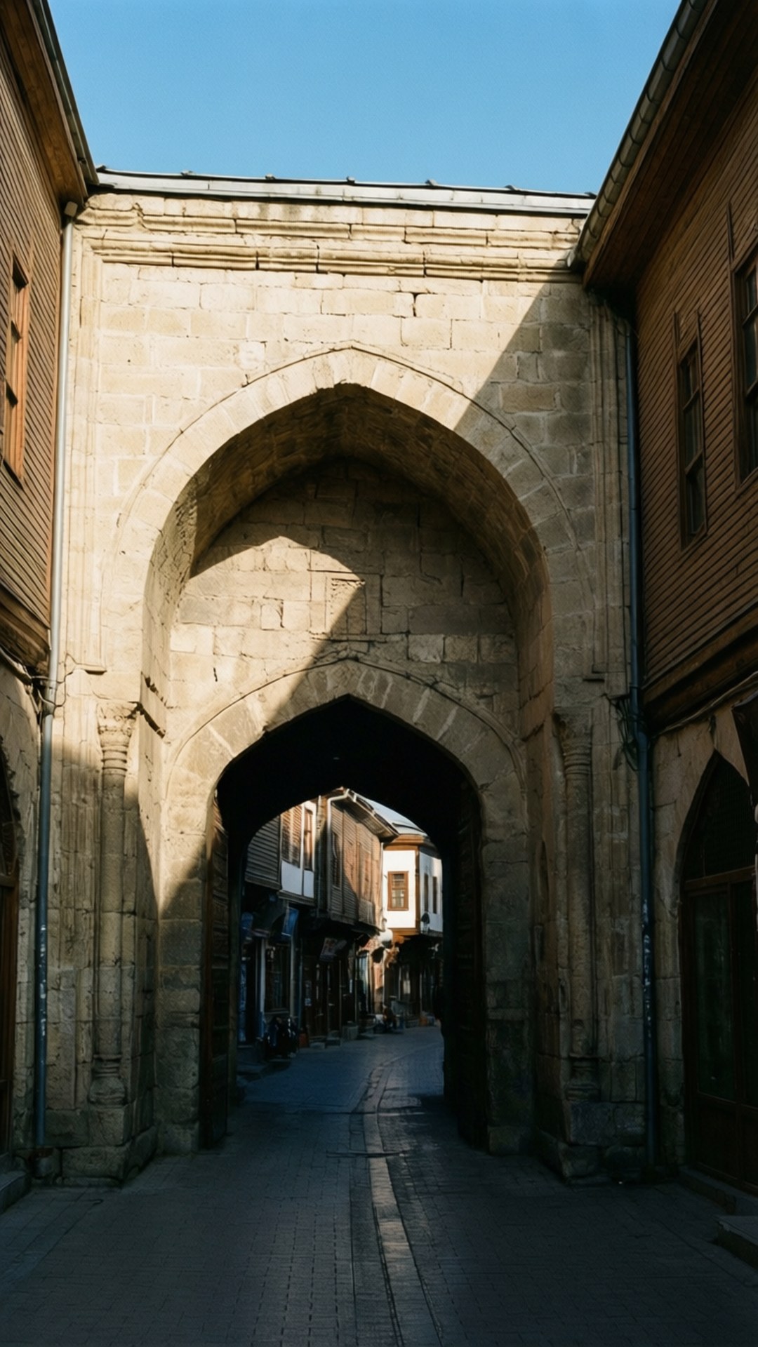 Kapu Cami Archway 壁紙 — 一個陽光照耀的石拱門框住了一條狹窄的鵝卵石街道，兩旁是歷史悠久的木建築，上方是清澈的藍天。