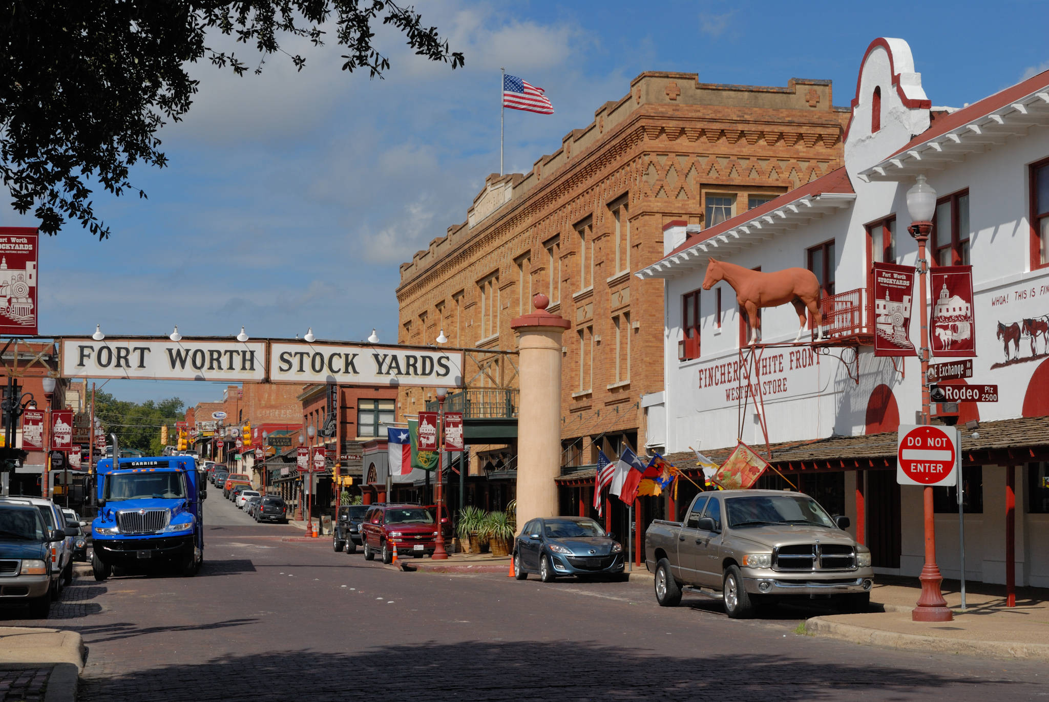 Worth 桌布 壁紙 — Fort Worth Stock Yards 一條陽光普照的街道，有歷史悠久的磚造建築、馬匹雕像和一輛藍色卡車停放在晴朗的藍天下。